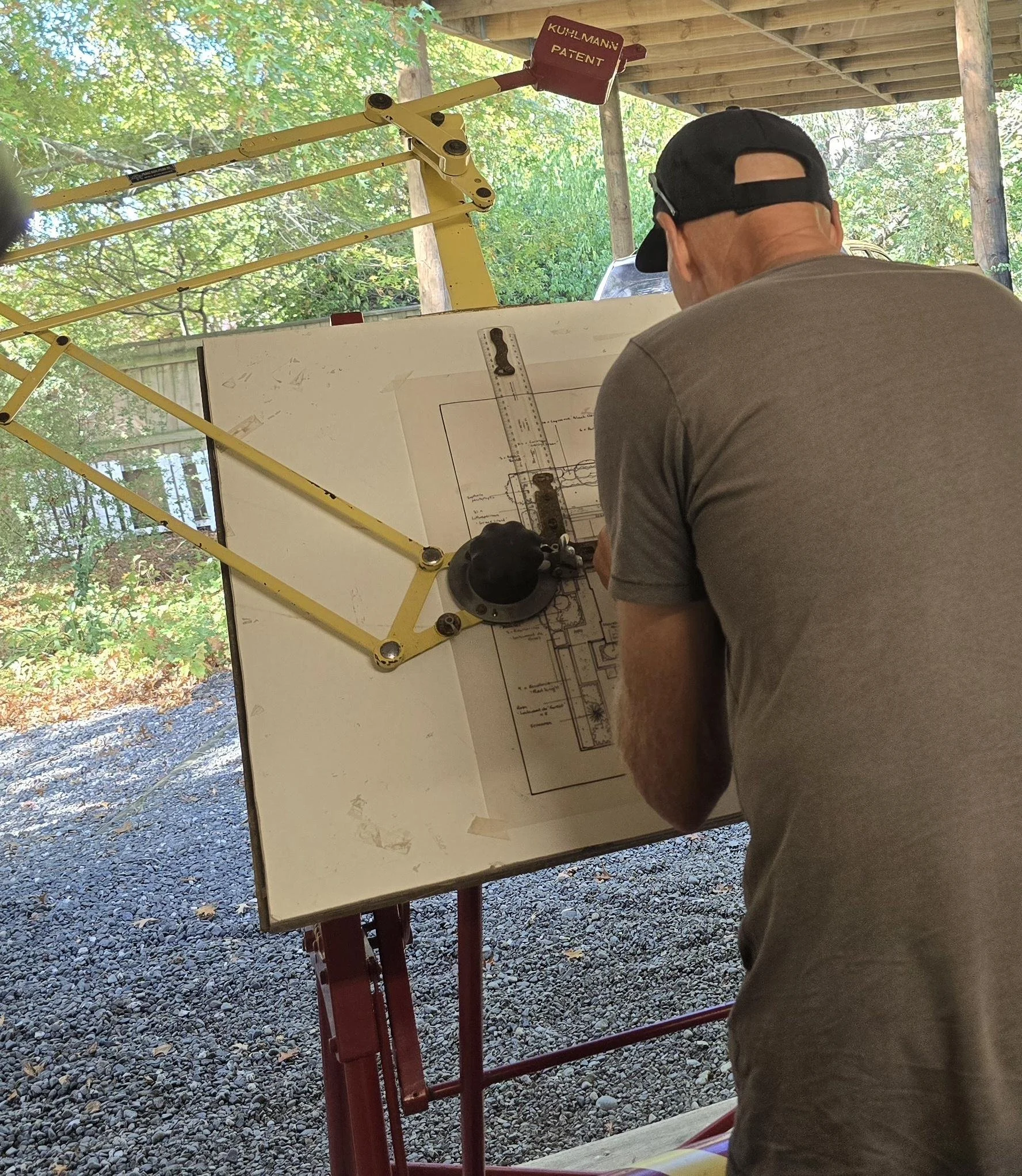 A man wearing a black cap and gray shirt, seated at an outdoor construction planning area, looking at a blueprint on an easel. The background includes green trees and a wooden roof.