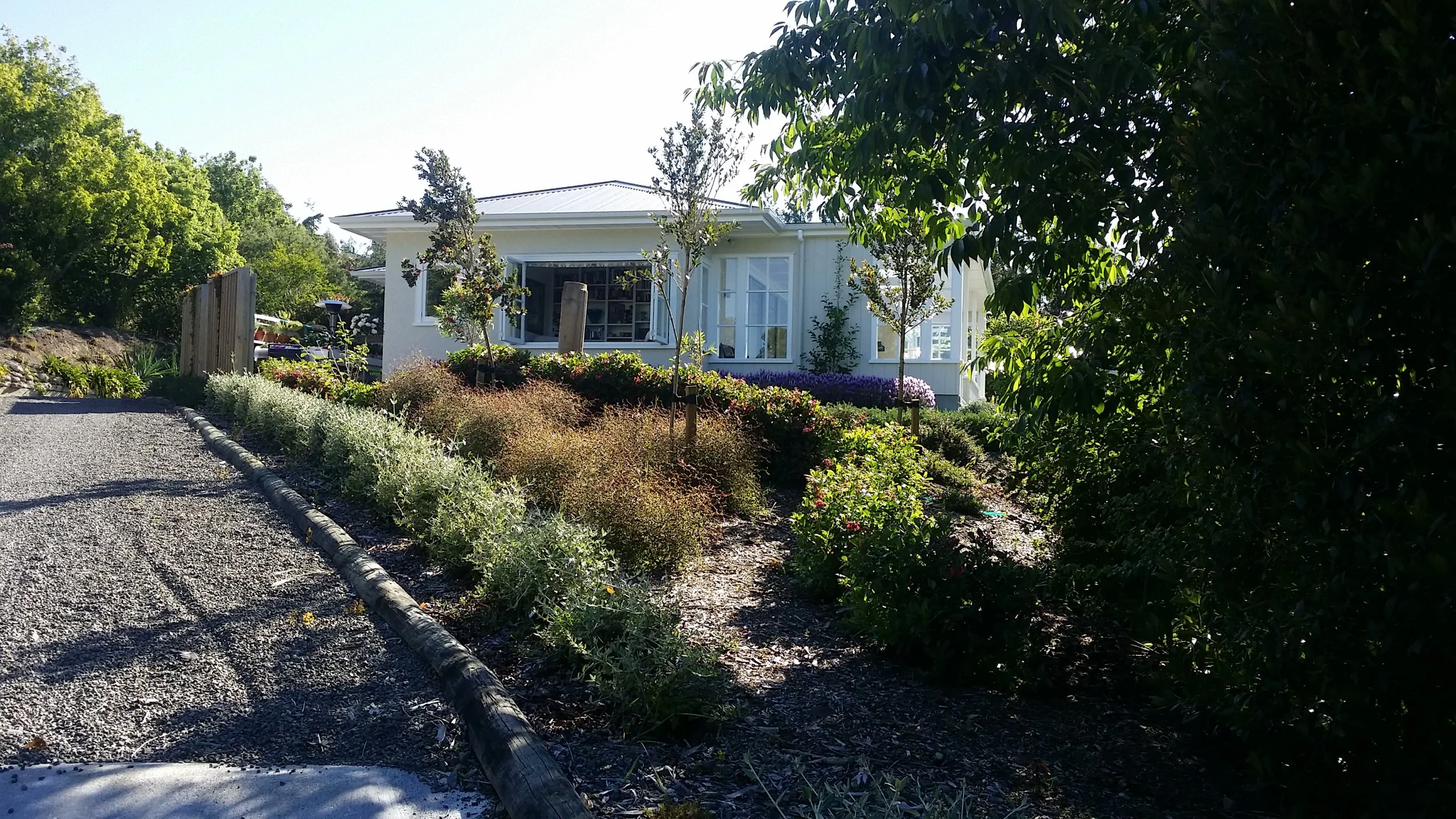 A white house surrounded by greenery and landscaped plants, with gravel path in the foreground and bright sunlight.