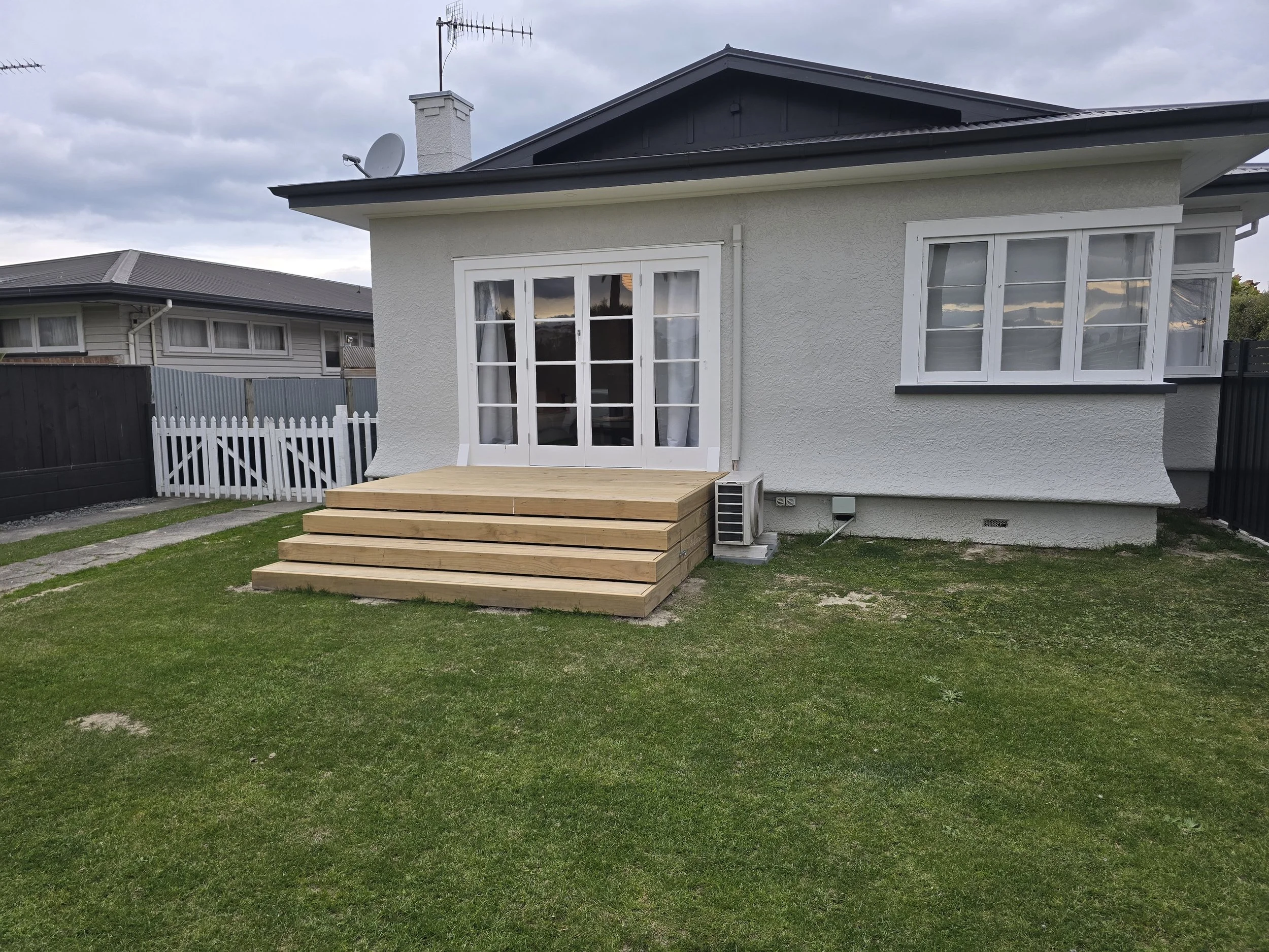 Backyard of a house with a small deck and a grassy lawn. The house has white walls, glass doors, and a window. There are two houses visible in the background with cloudy skies overhead.