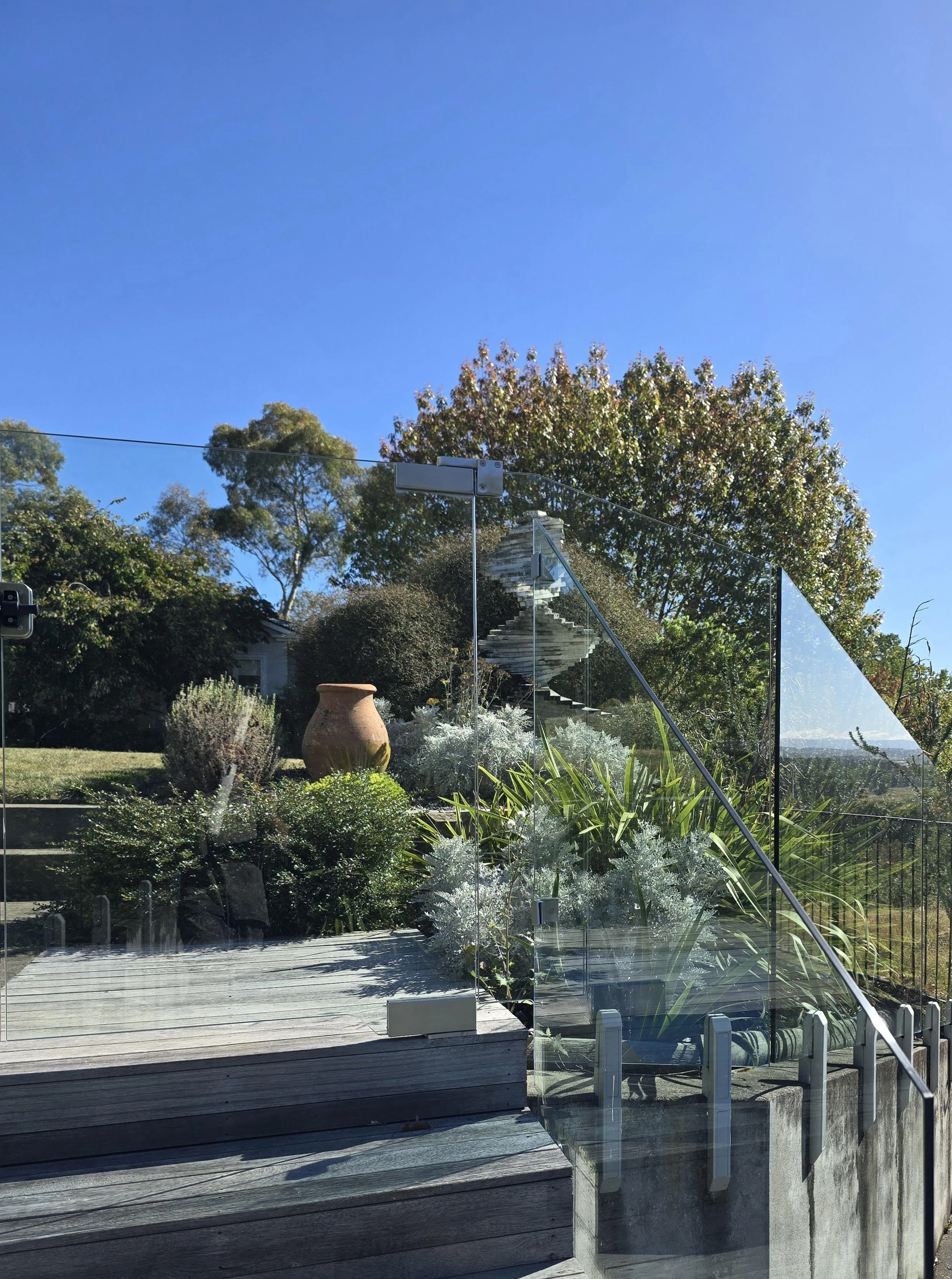 A modern outdoor space with wooden steps, a glass railing, and a lush garden with plants, trees, and a large clay pot under a clear blue sky.