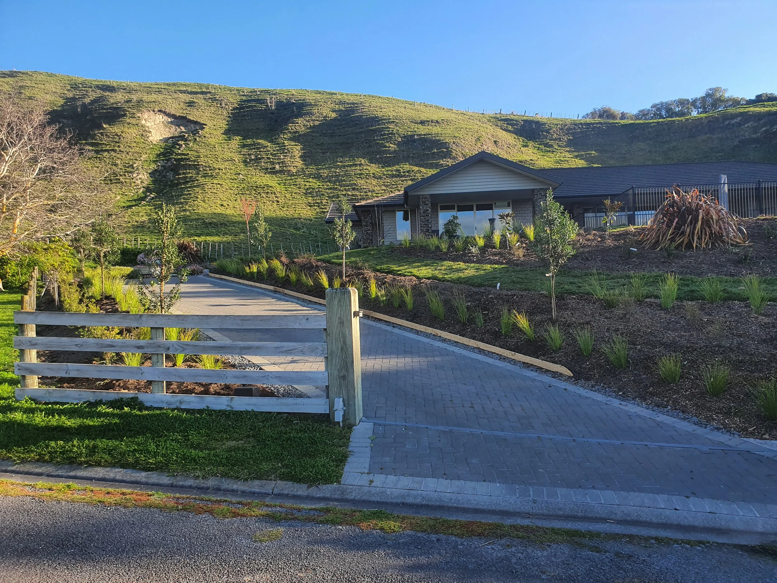 A house with a dark roof and large windows on a hillside with green grass and plants. A paved path runs in front of the house, and a wooden fence is at the edge of the sidewalk. In the background, green hills and trees under a clear blue sky are visi