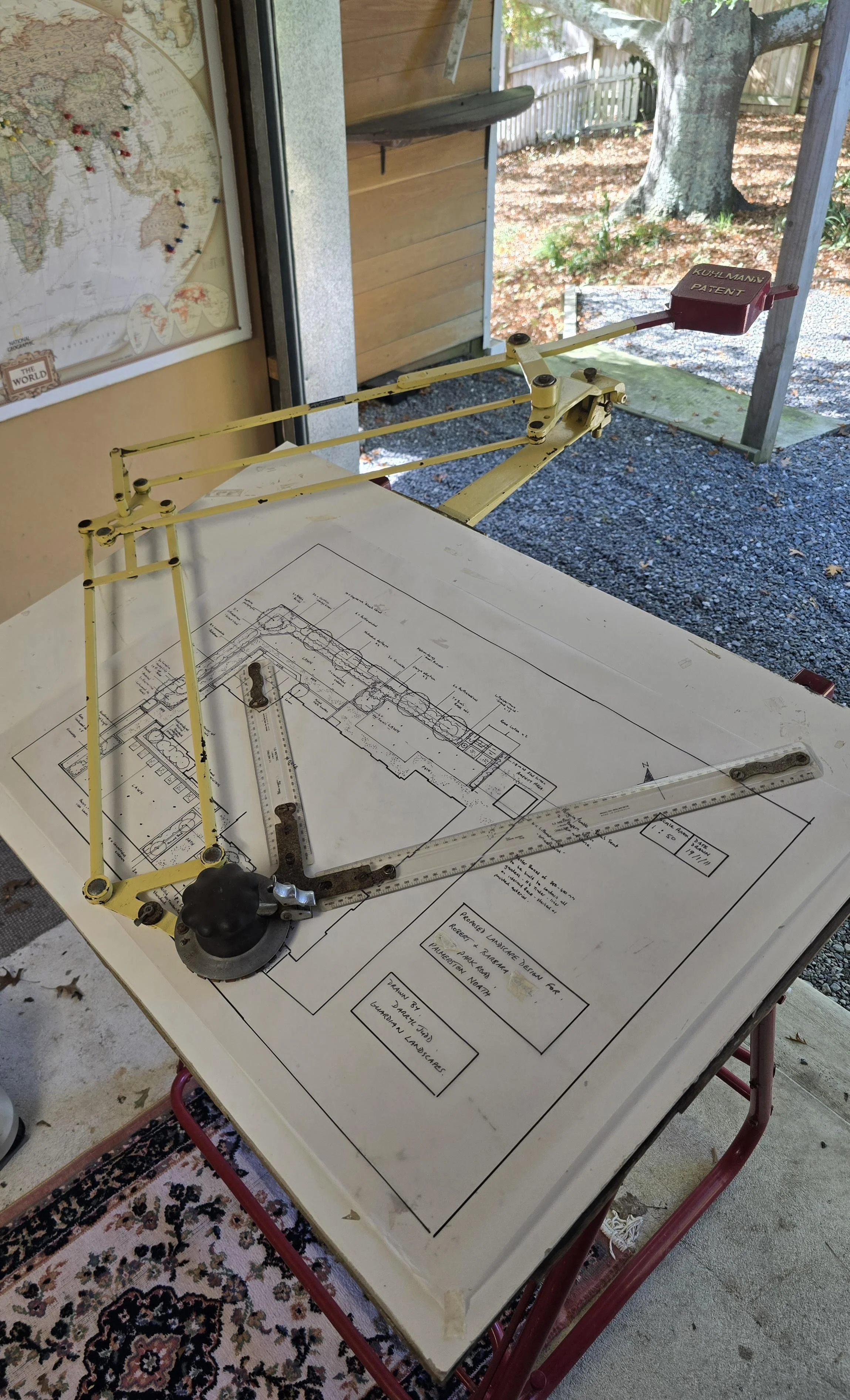 Architectural blueprints on a table with a draft person's drafting machine, in an indoor and outdoor setting, with a map pinned on the wall, a tree visible outside, and a patterned rug on the floor.