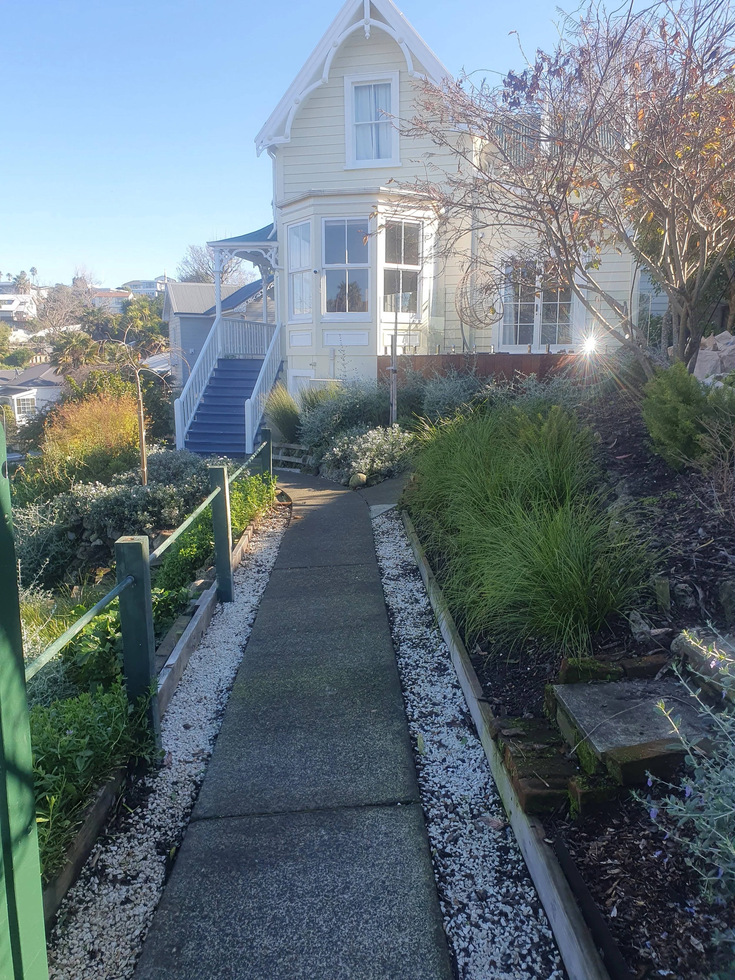 A curved sidewalk leads to a white house with a staircase and a large bay window, flanked by plants and trees, in a residential neighborhood on a sunny day.