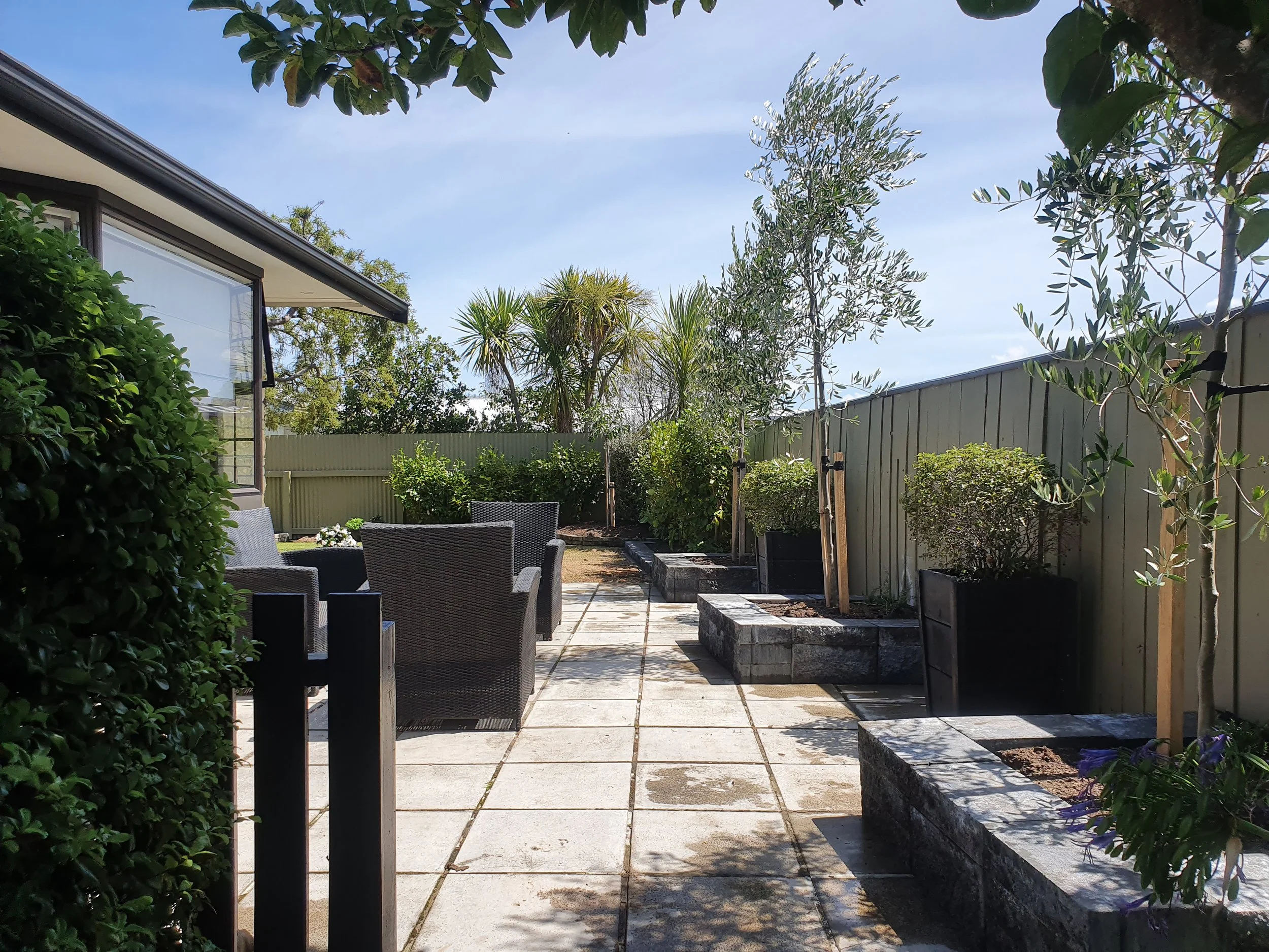 A backyard patio area with outdoor furniture, trees, and a fence under a clear blue sky.