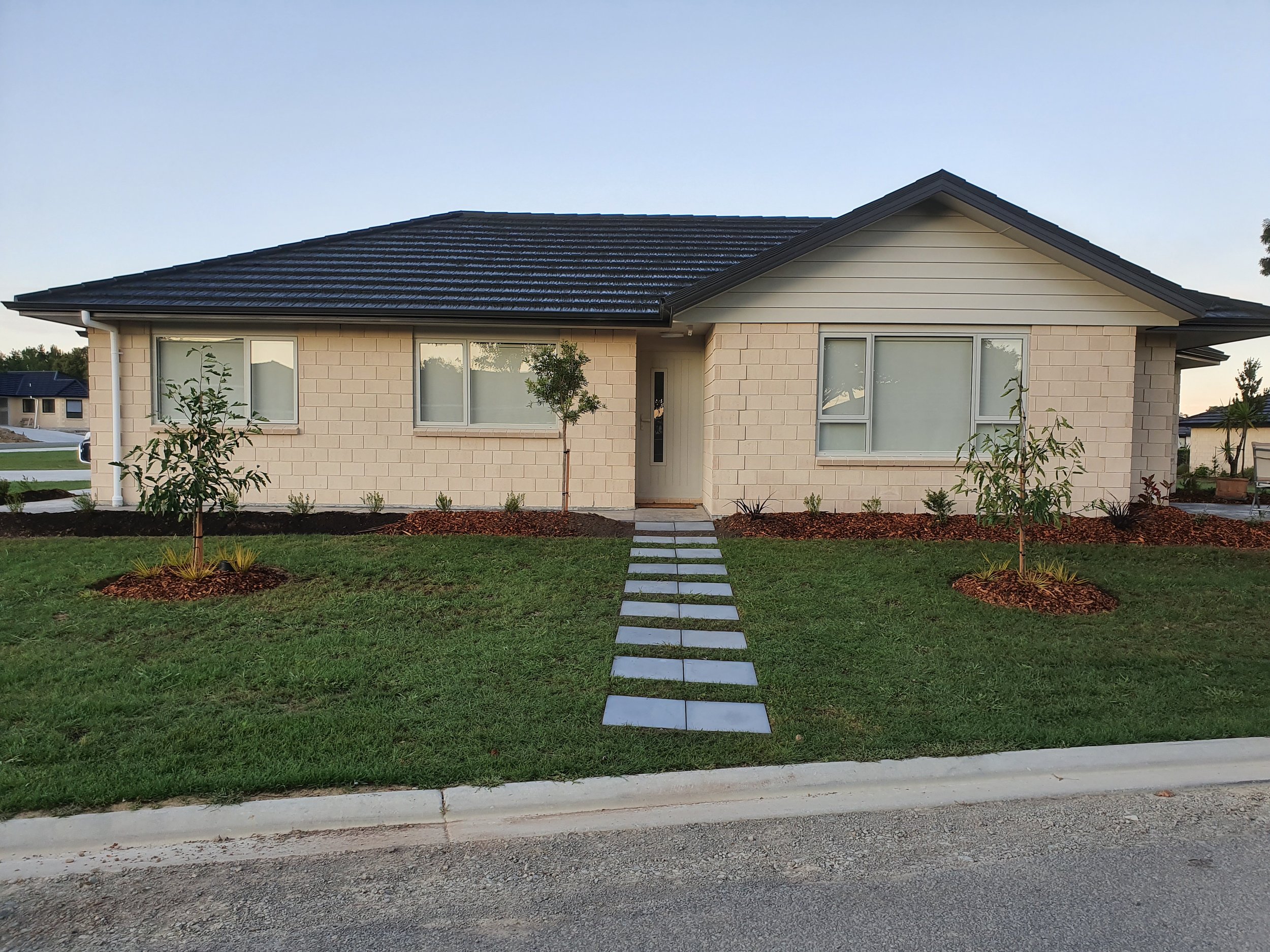 A modern single-story house with a black tiled roof, beige brick exterior, two large windows, a narrow front door, small trees and plants in the front yard, and a pathway leading to the entrance, with other houses visible in the background during day