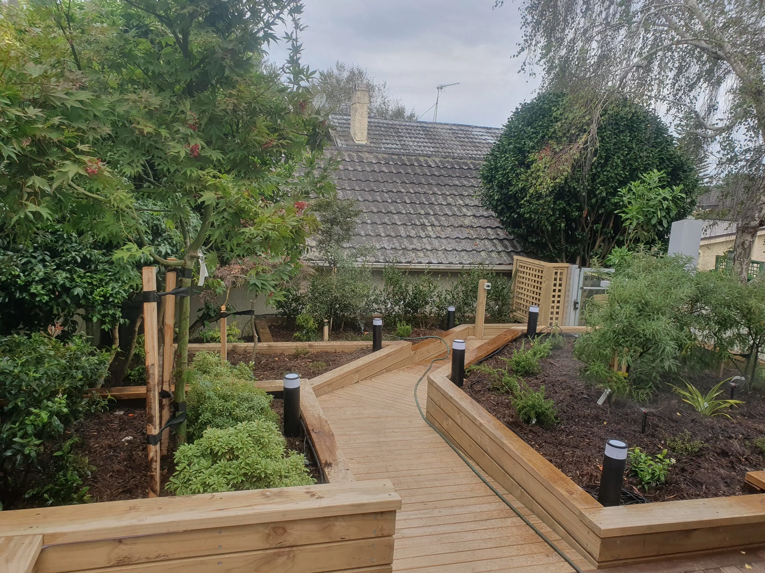 A backyard garden area with a wooden walkway, surrounded by green plants and trees. There is a house with a gray tiled roof in the background and a cloudy sky overhead.