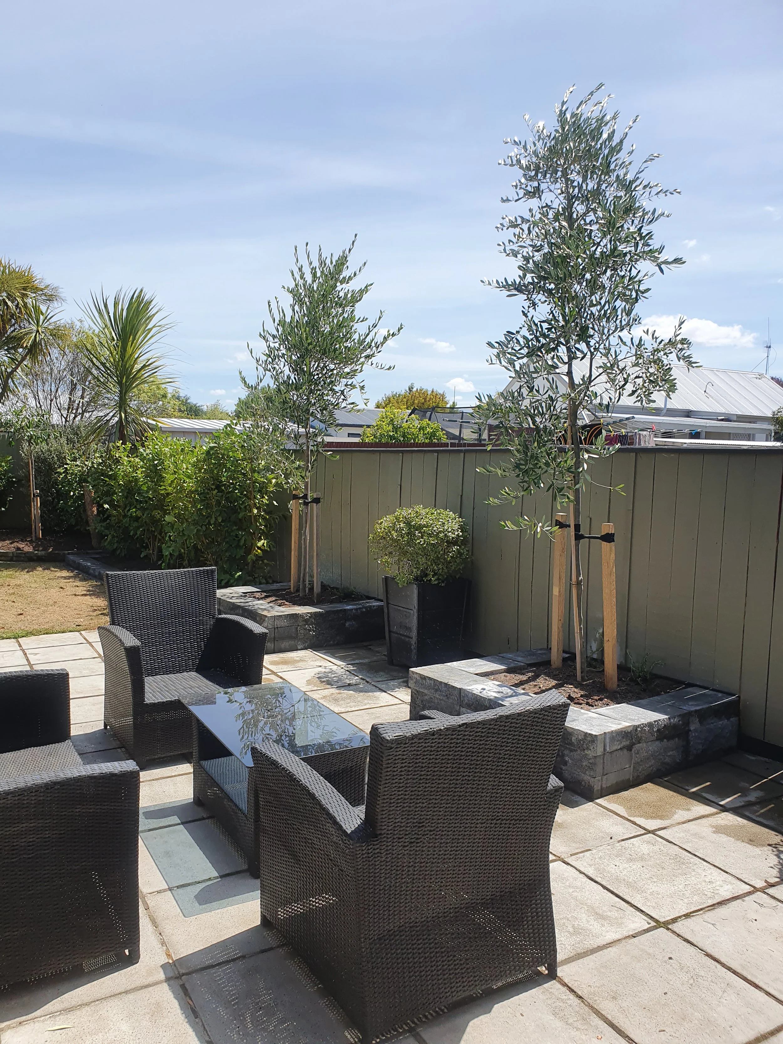 An outdoor patio area with black wicker chairs and a glass-top table, surrounded by a garden with various trees and plants, and a grey fence in the background under a partly cloudy sky.
