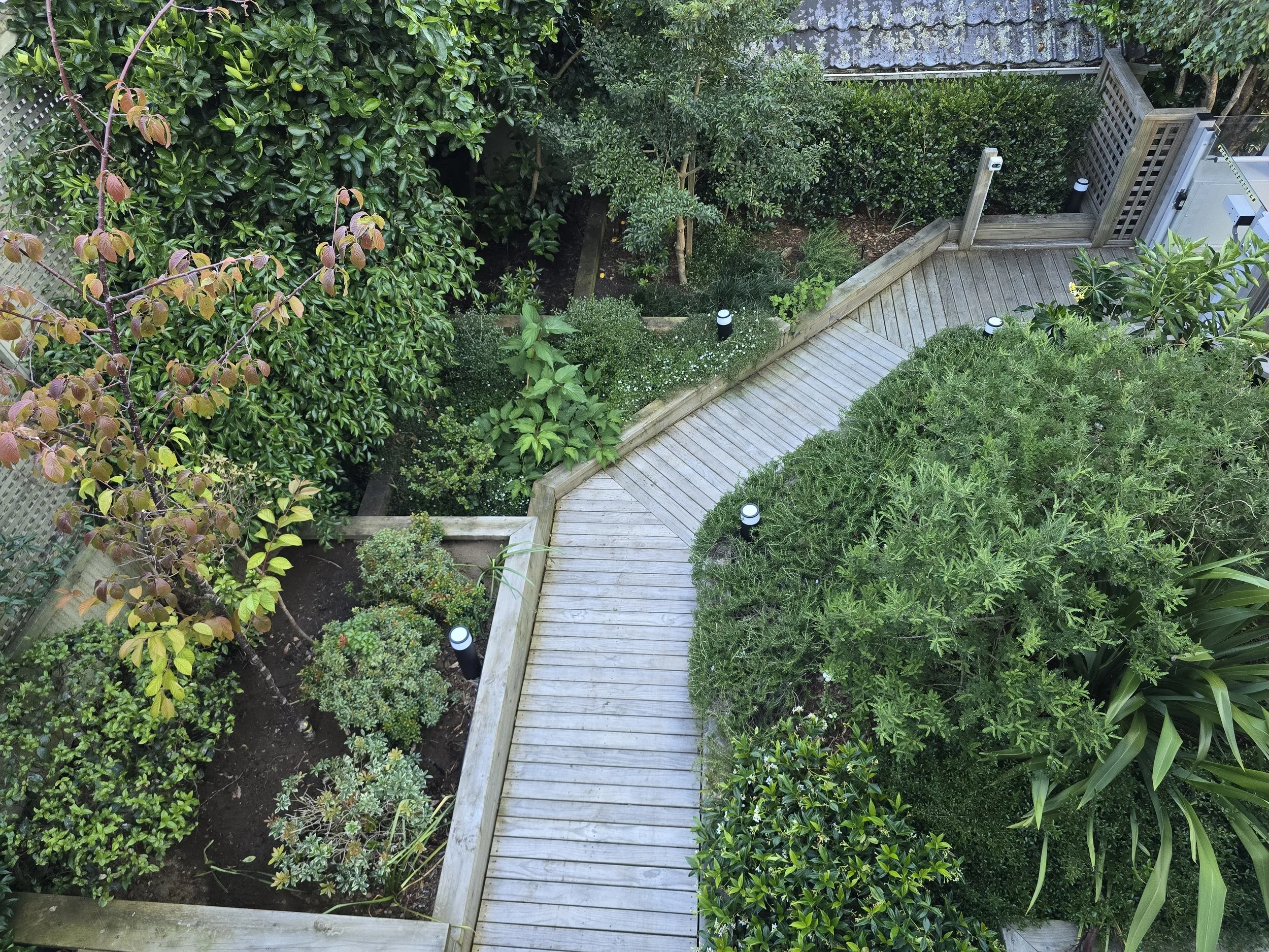 Overhead view of a backyard garden with a wooden deck, lush green bushes, trees, and a small garden bed with plants and flowers.