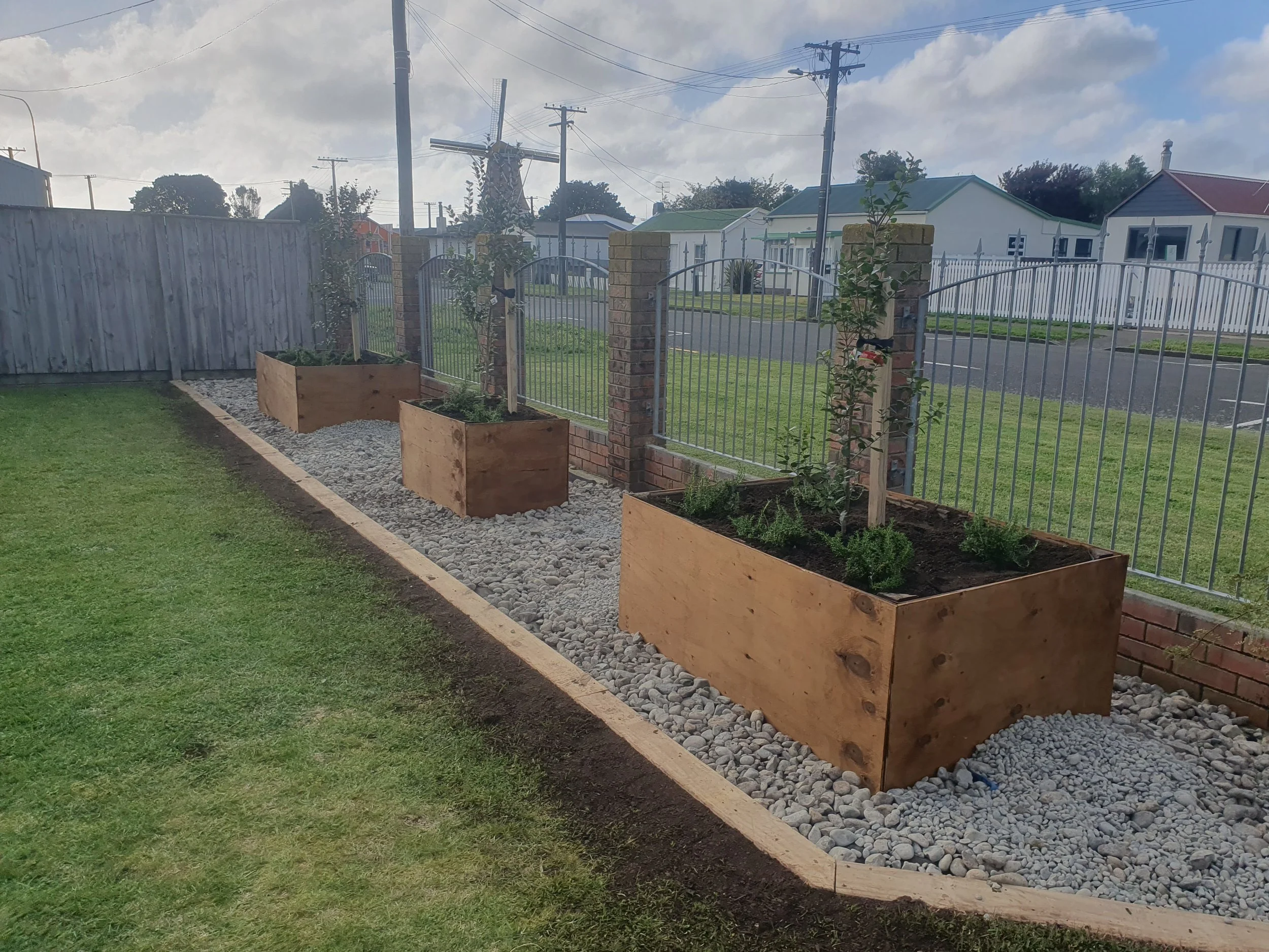 A backyard with a garden bed, wooden planters, and a gravel border, enclosed by a metal fence and brick columns. There are some small trees and plants growing in the planters. The background shows a street with houses, power lines, and a windmill str