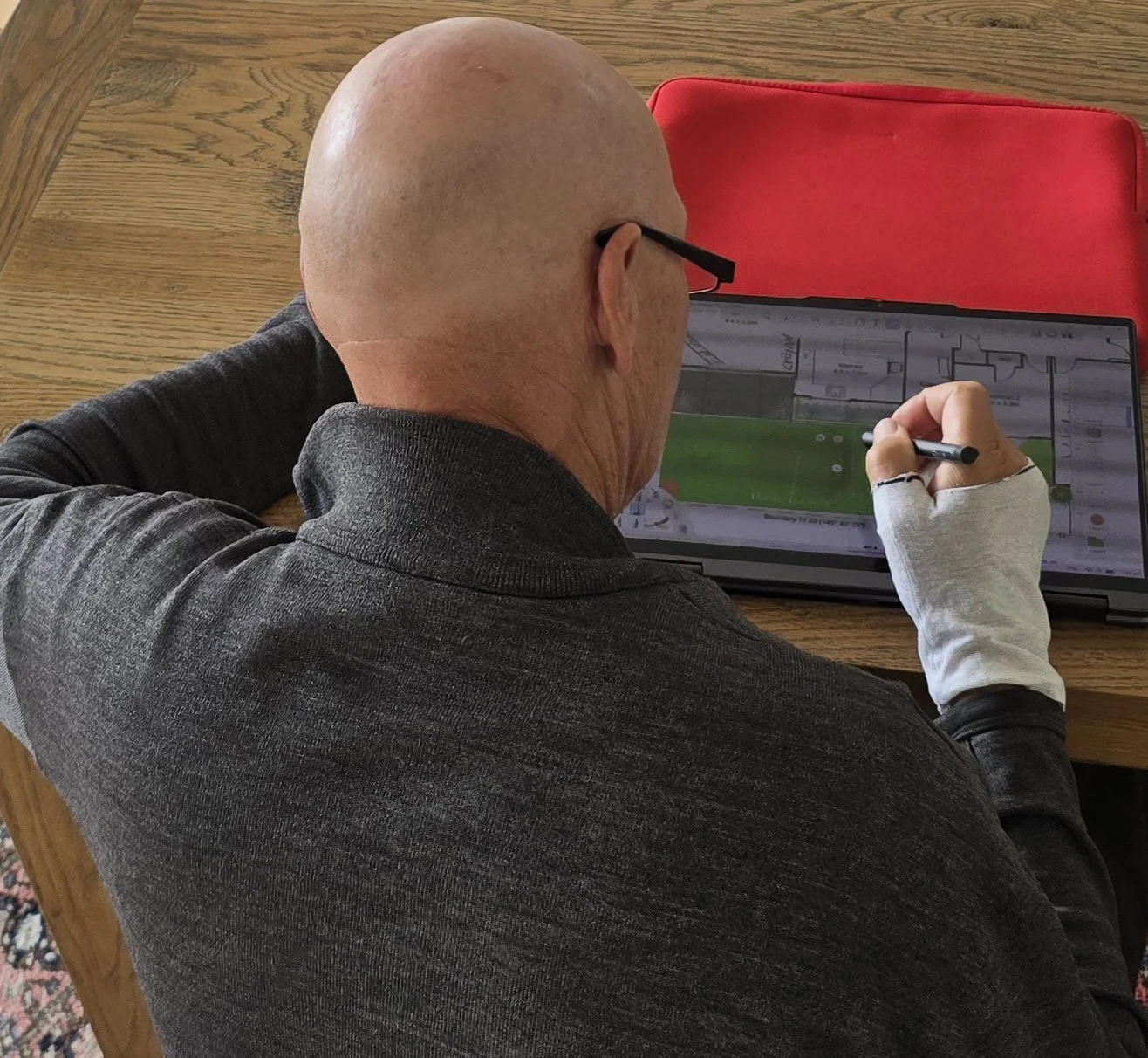A bald man with glasses, wearing a dark gray shirt and a white sleeve, is sitting at a wooden table, working on a digital device with a stylus. A red bag is nearby.