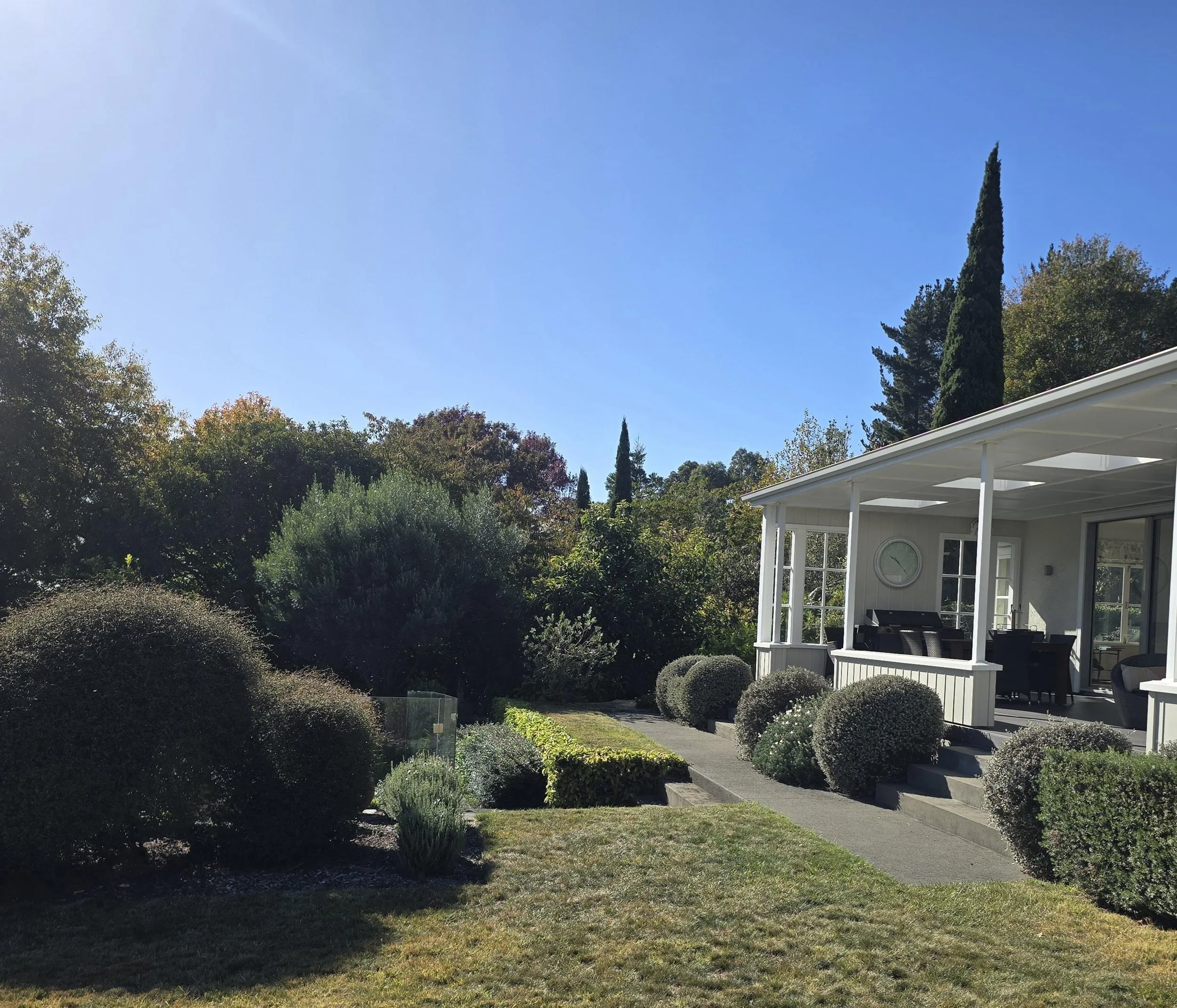 A backyard garden scene with a white house featuring a porch, surrounded by various bushes and trees, under a clear blue sky.