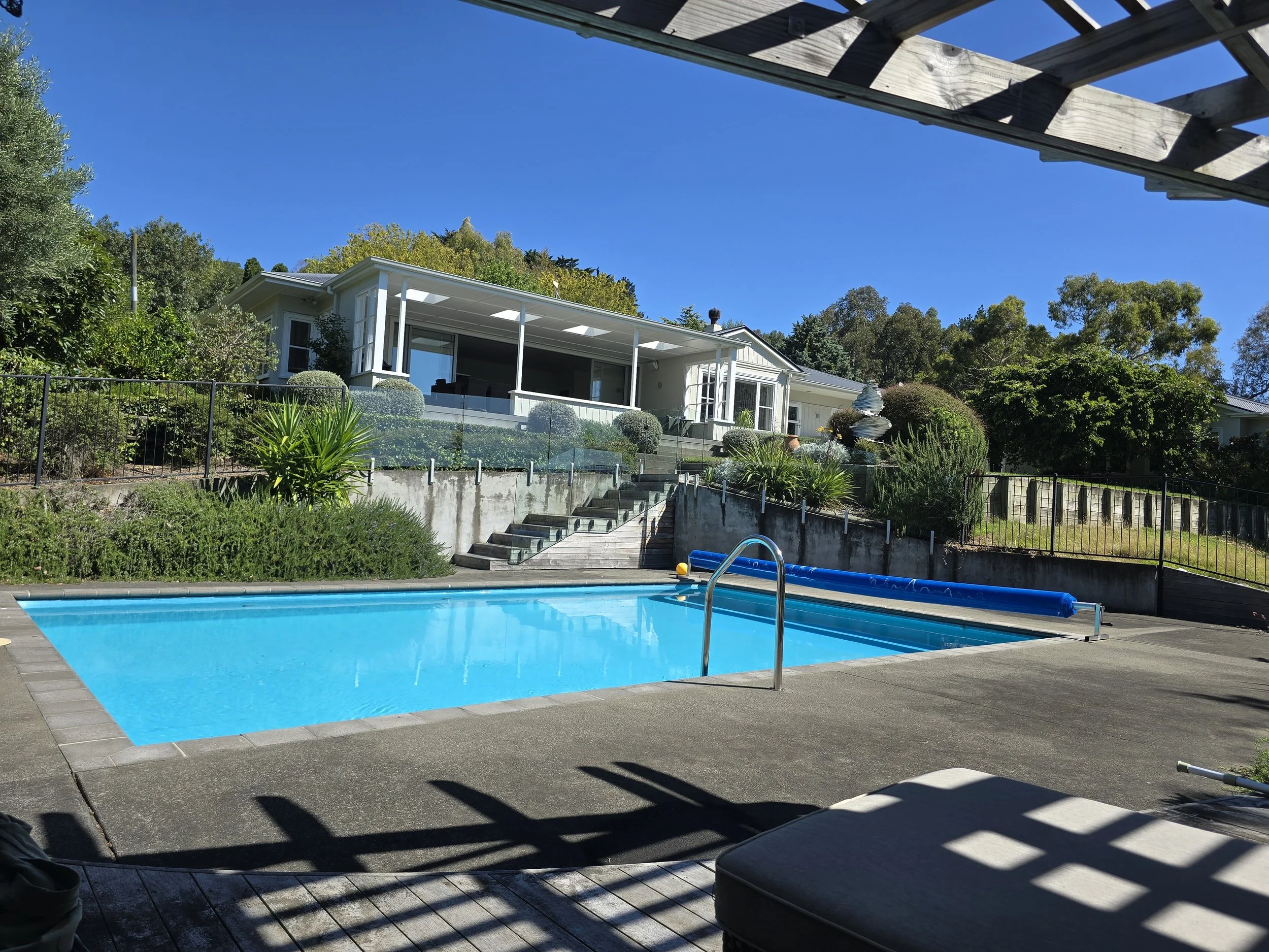 View of a backyard with a swimming pool, a house with a shaded porch, stairs leading up to the house, lush green trees, and a wooden deck with a pool cover rolled up nearby.