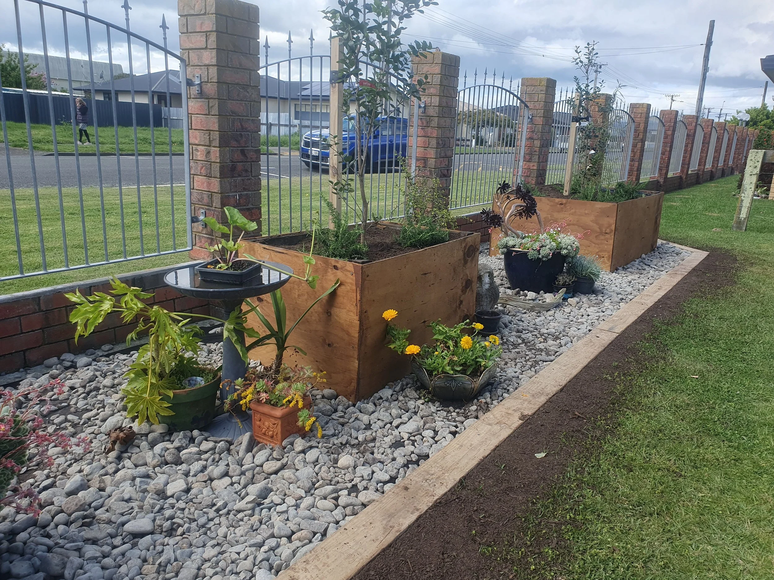 A garden area with large wooden planters filled with trees and flowering plants, surrounded by a layer of white pebbles. A brick wall and metal gate enclosure surrounds the garden, and beyond are a street, parked cars, and a pedestrian walking on the