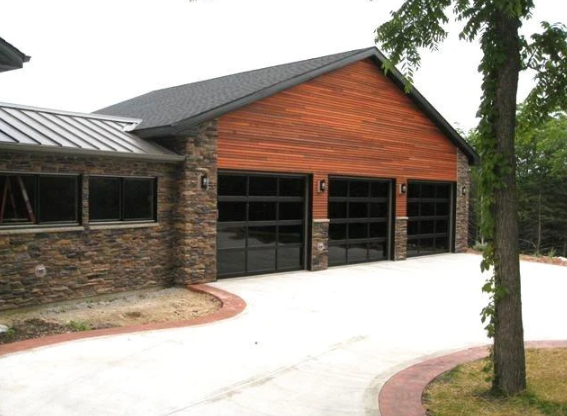 Modern house with a wooden and stone exterior, three garage doors, and a curved driveway surrounded by grass and trees.