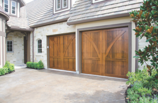 Two wooden garage doors in front of a stone house with manicured bushes and a driveway.
