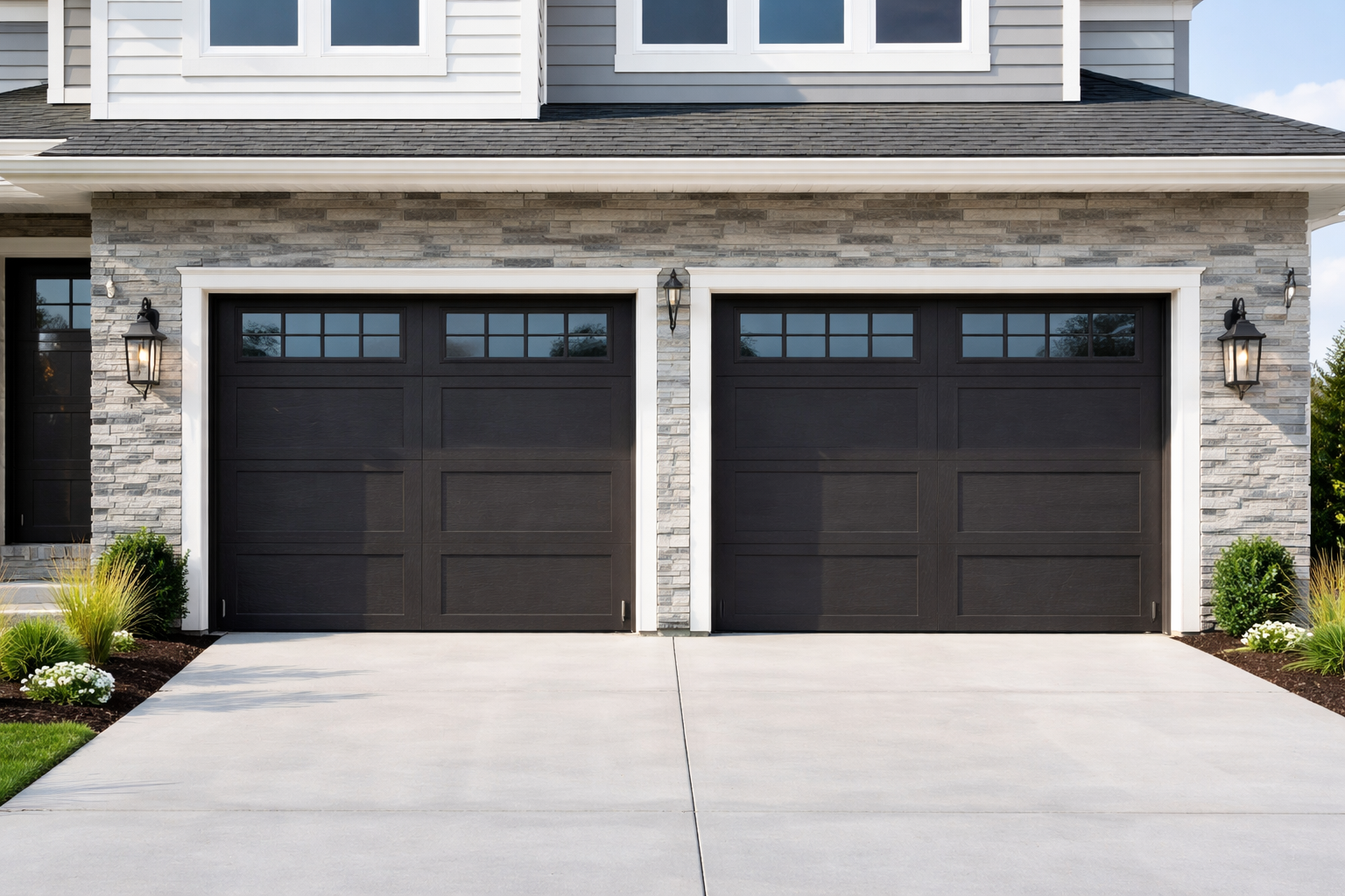 Front view of a modern house with two black garage doors, stone facade, two wall-mounted lanterns, landscaping with bushes, and a concrete driveway.