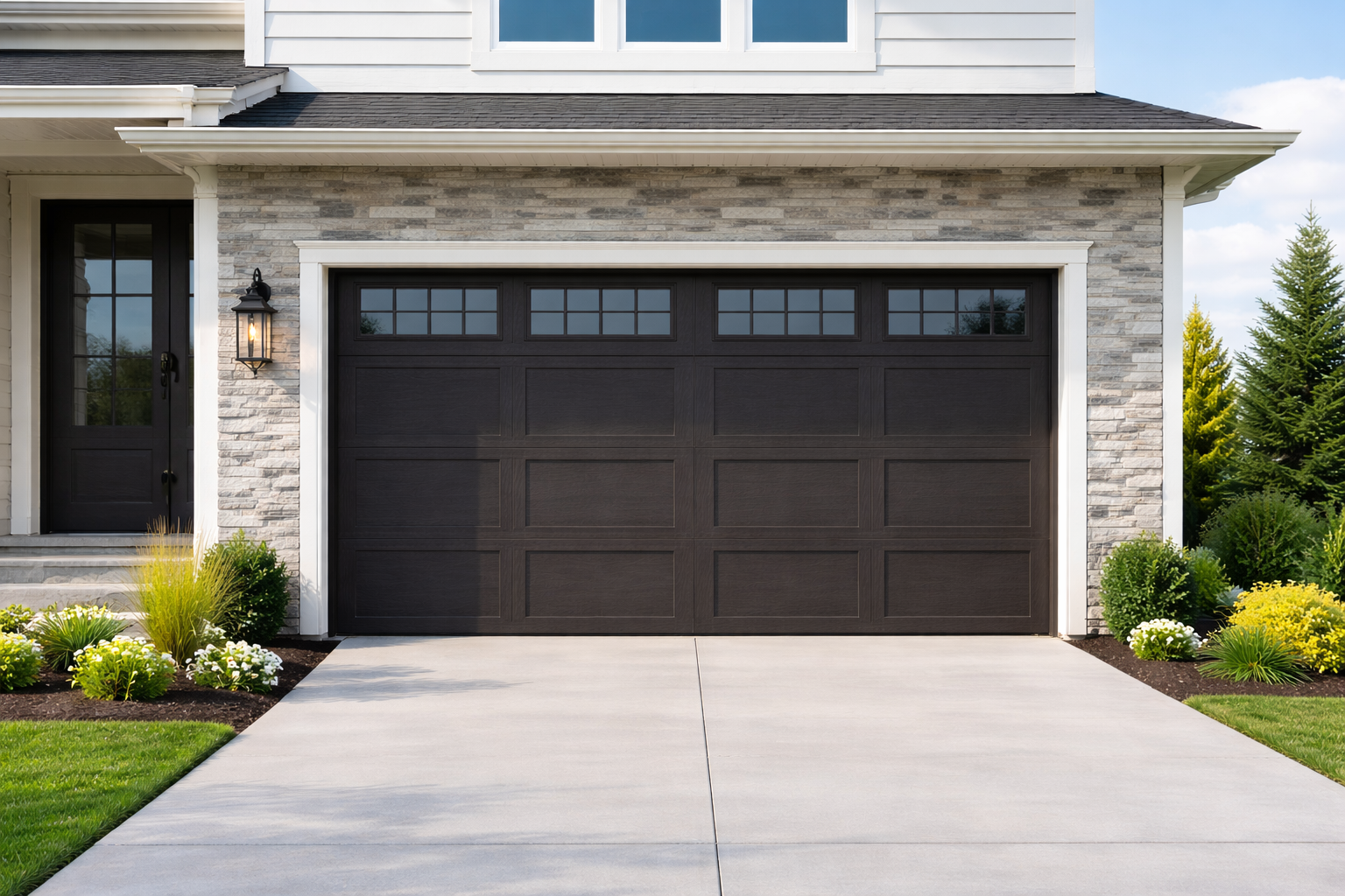 Front view of a modern house garage with black paneled garage door, side wall lamp, and flowerbed with white and yellow flowers and green bushes.