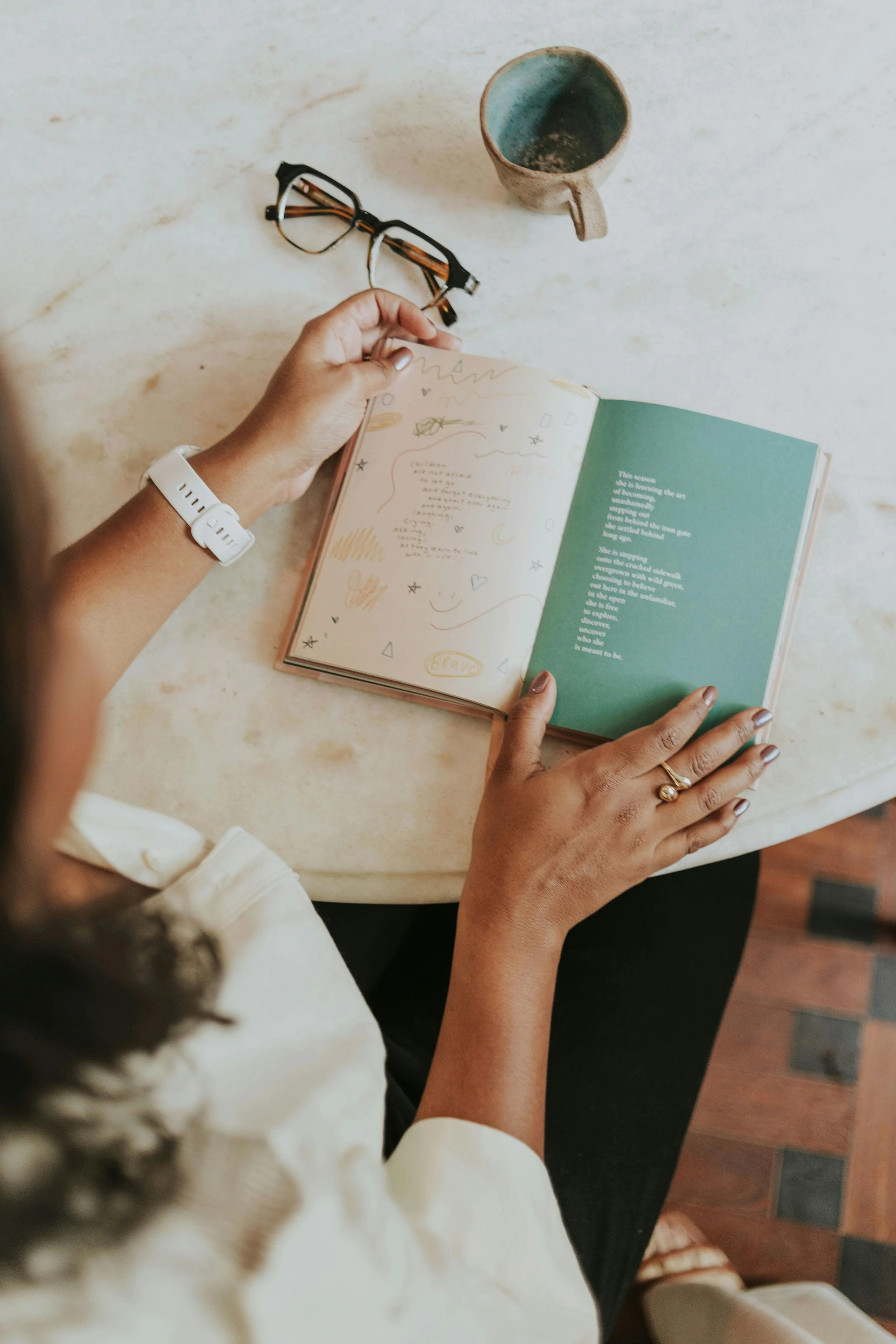 A person with a white watch on their wrist, reading a colorful, handwritten journal. There is a ceramic mug with a handle and a pair of glasses on a white marble table.