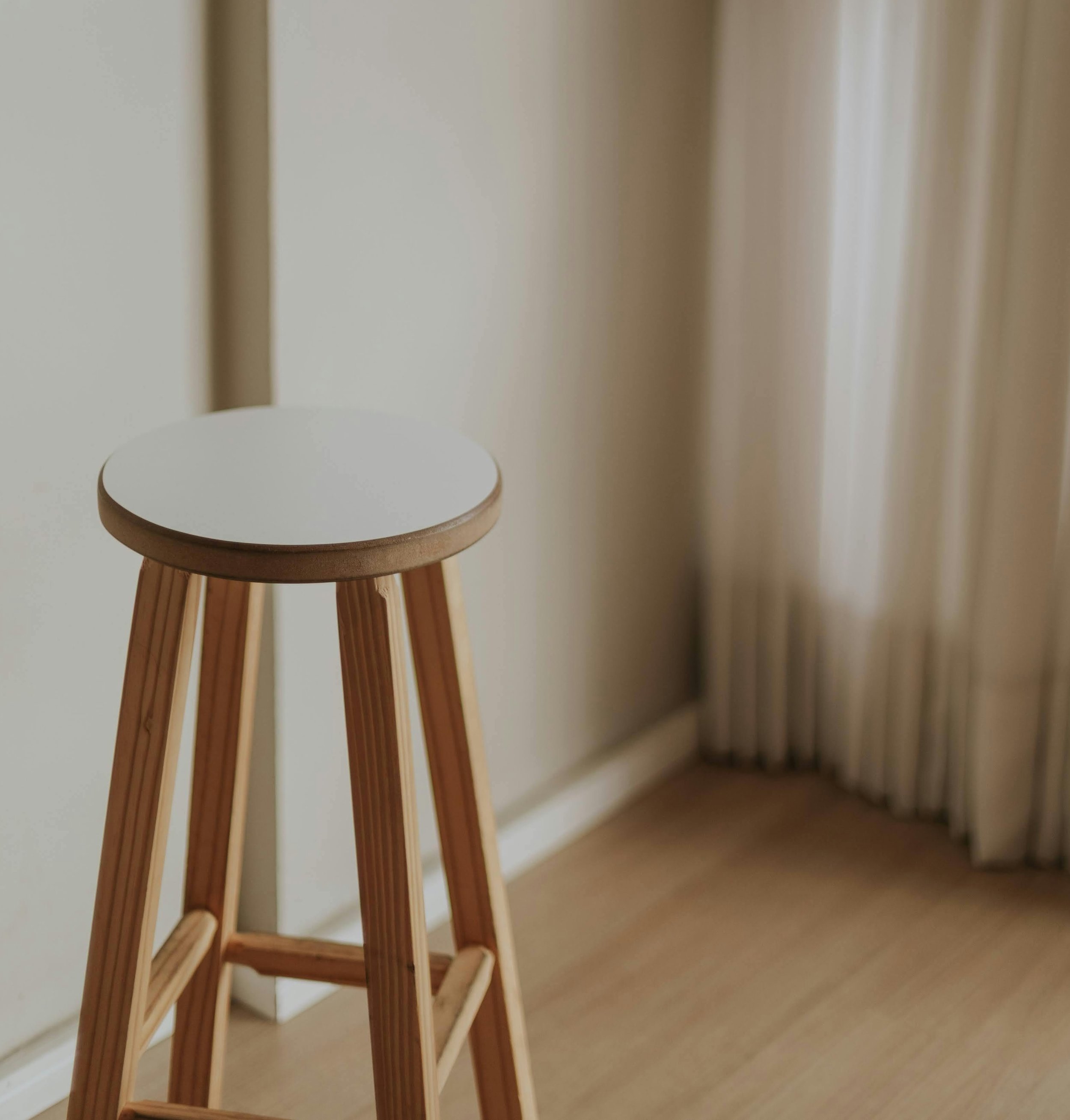 A small wooden stool with a round white seat in front of beige curtains on a wooden floor.