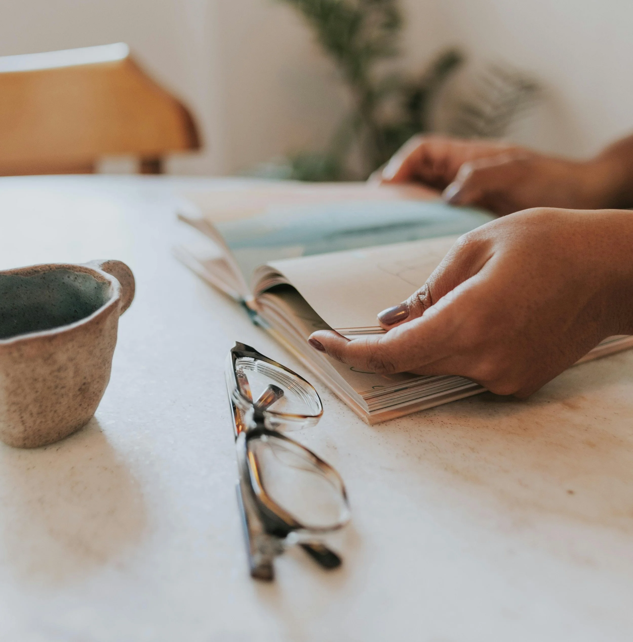 Close-up of a person’s hands holding a pen and writing in a notebook on a white table with a coffee mug nearby, and glasses resting on the table.