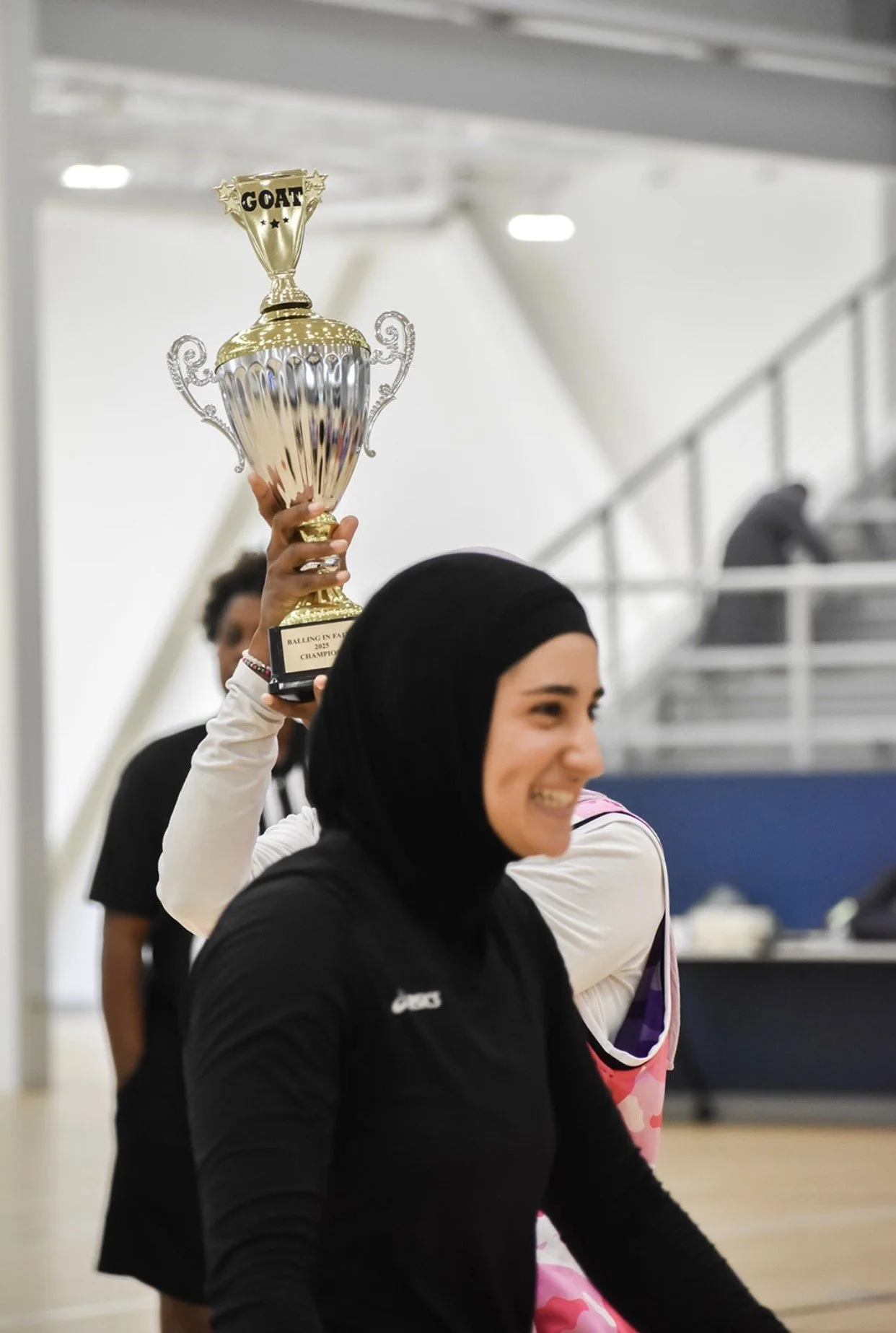 A woman wearing a black hijab and athletic clothing smiling, holding a trophy above her head, in an indoor sports venue.