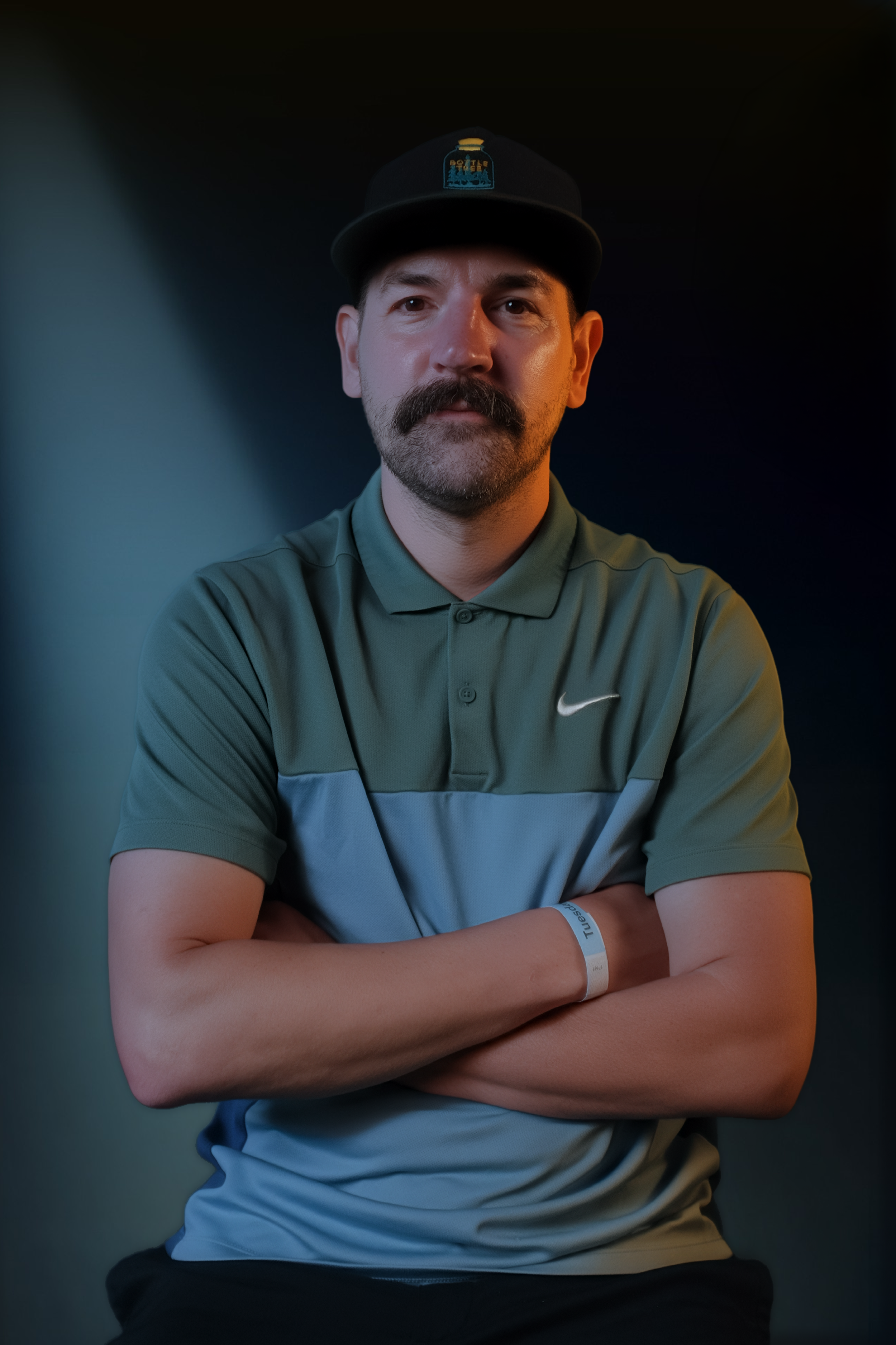 Man with a beard and mustache wearing a cap and a polo shirt with his arms crossed, standing against a dark background.