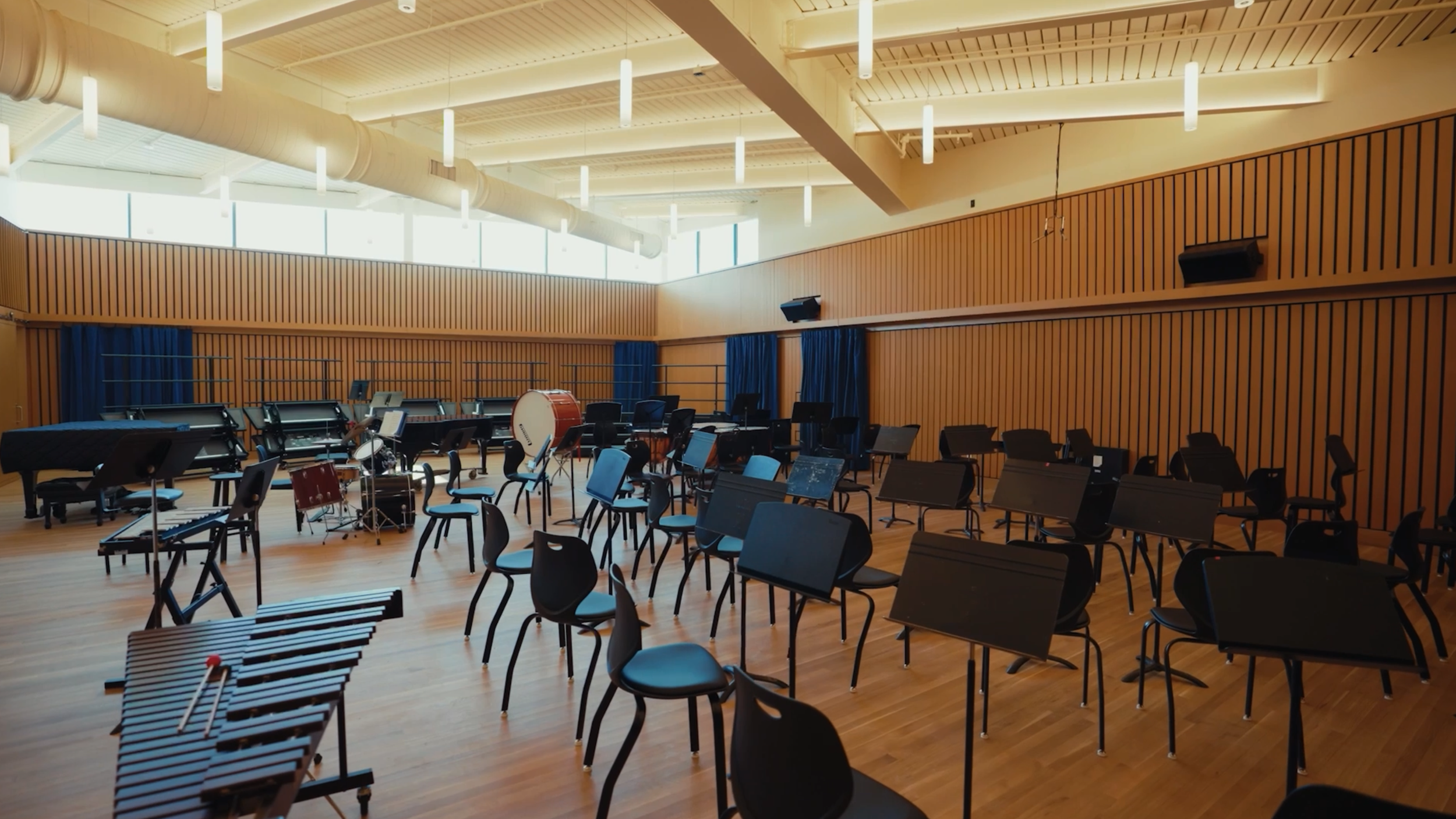 Empty music rehearsal room with black chairs, music stands, percussion instruments, and large windows, wood-paneled walls, and modern lighting.