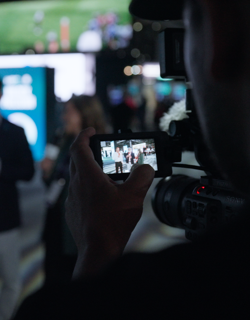 Person filming a presentation or event at an indoor venue with a professional camera. The screen of the camera shows a clearer view of speakers standing in front of a blue backdrop.