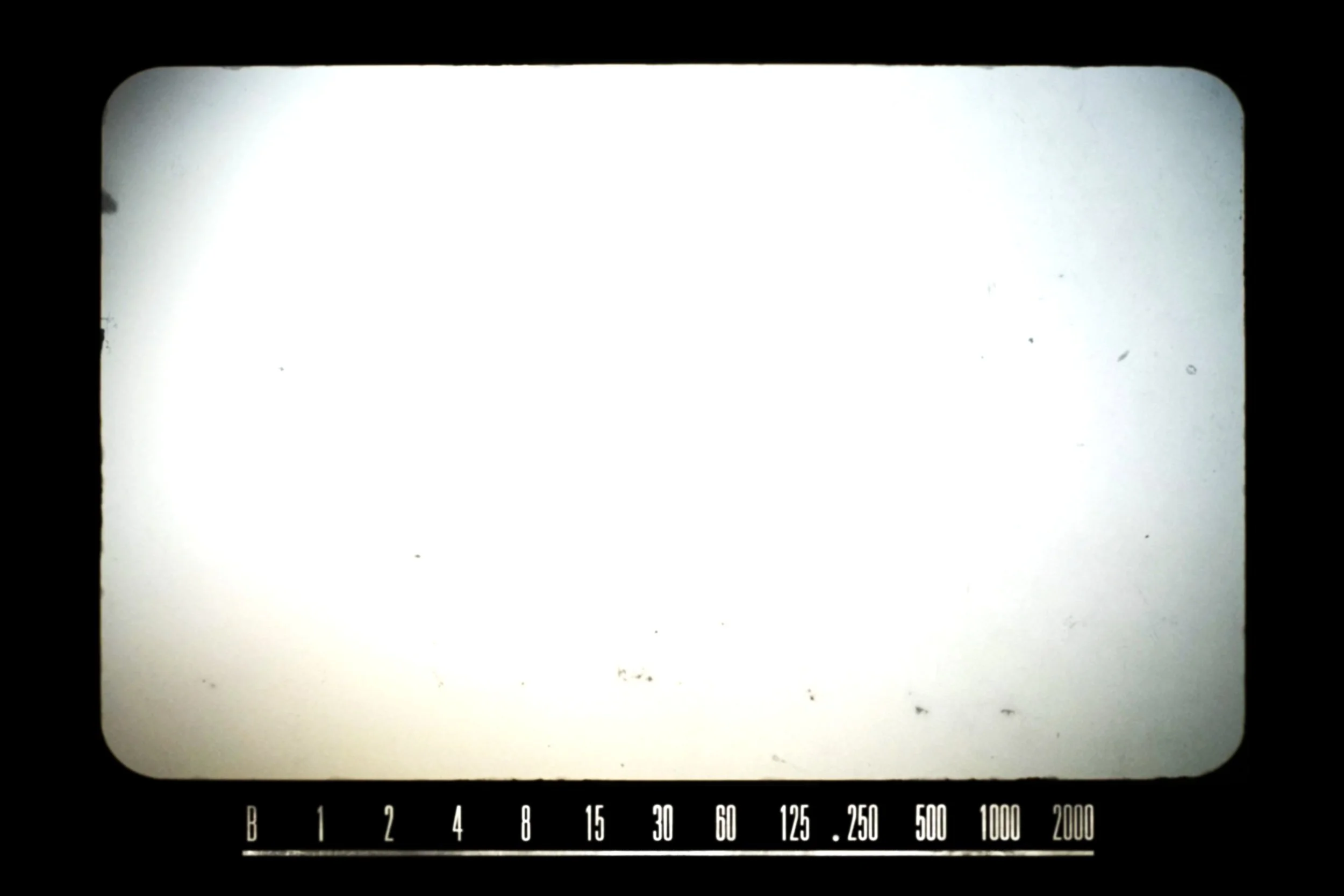 A black and white photo of a film camera screen showing a blank white frame with film exposure measurements at the bottom.