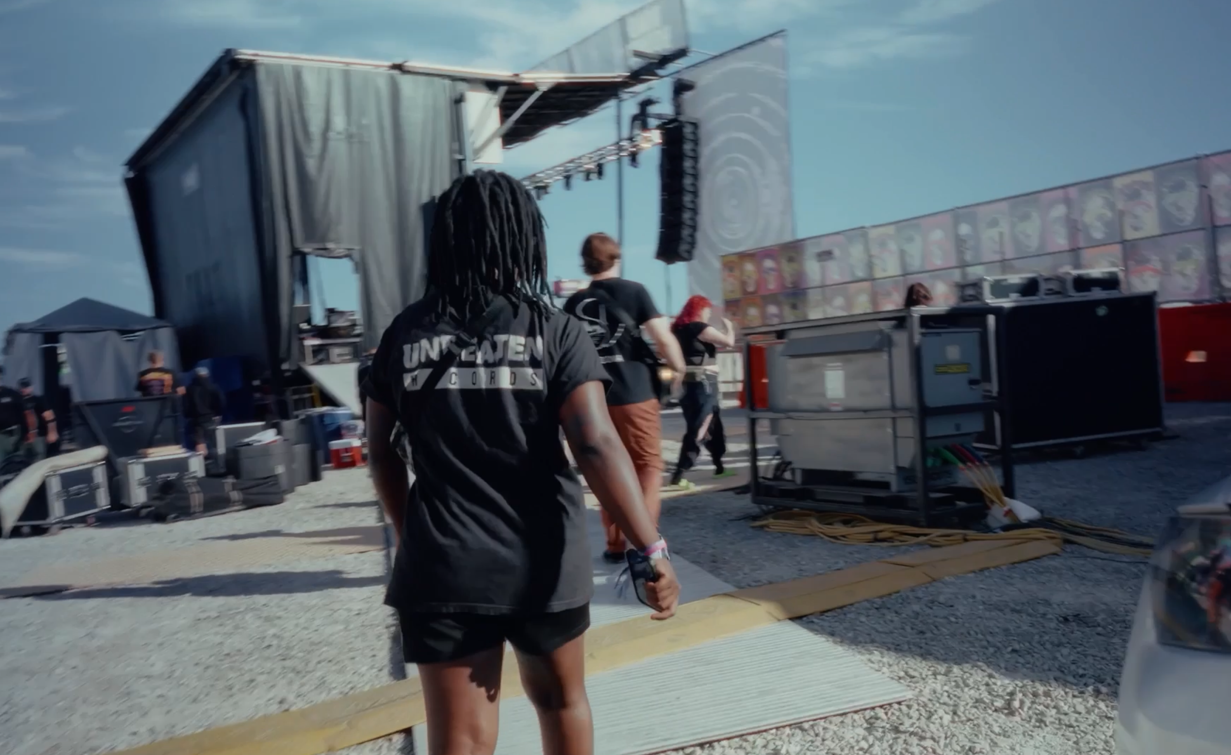 Back view of a person with long dark dreadlocks wearing a black t-shirt with white text that says 'UNCREATED' walking toward a stage at an outdoor event. There are other people nearby, equipment, and a large screen in the background under a partly cl