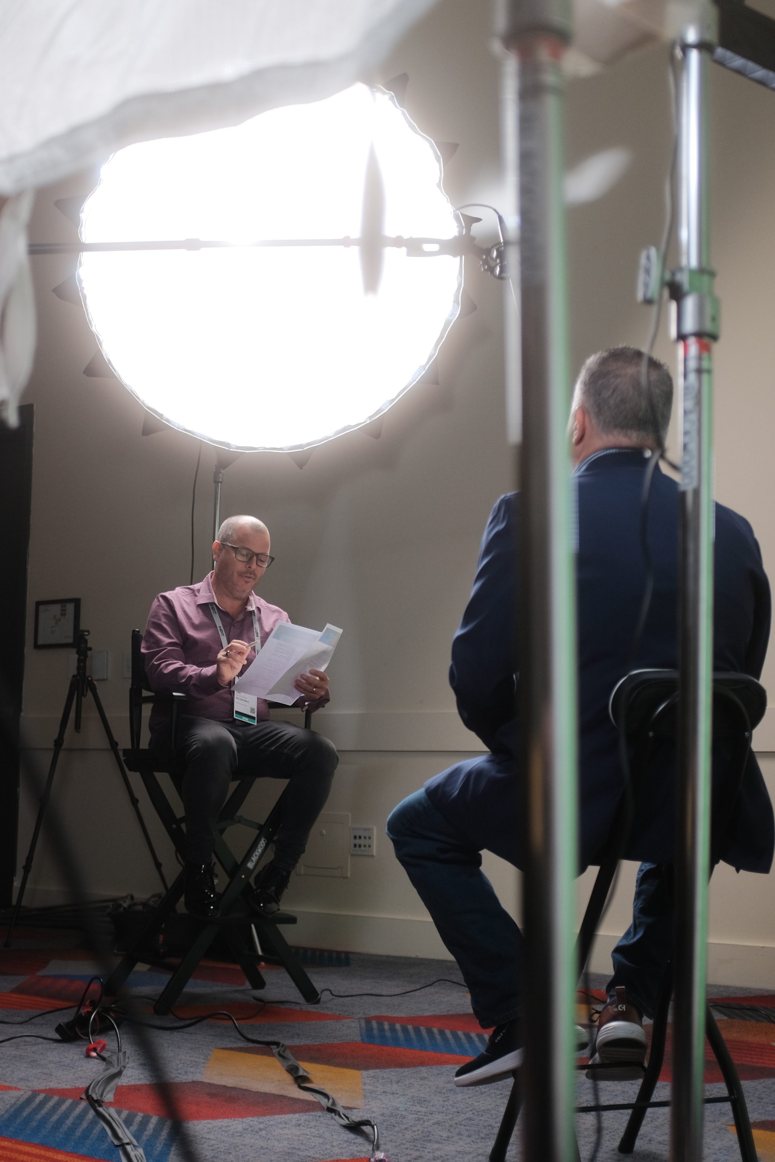 Two men are participating in a video interview or recording in a studio setting. One man is sitting on a stool, reading from a paper, while the other man, seen from the back, is seated and facing the first man. A bright light is overhead, and there is professional equipment, including tripods and cables, in the room.