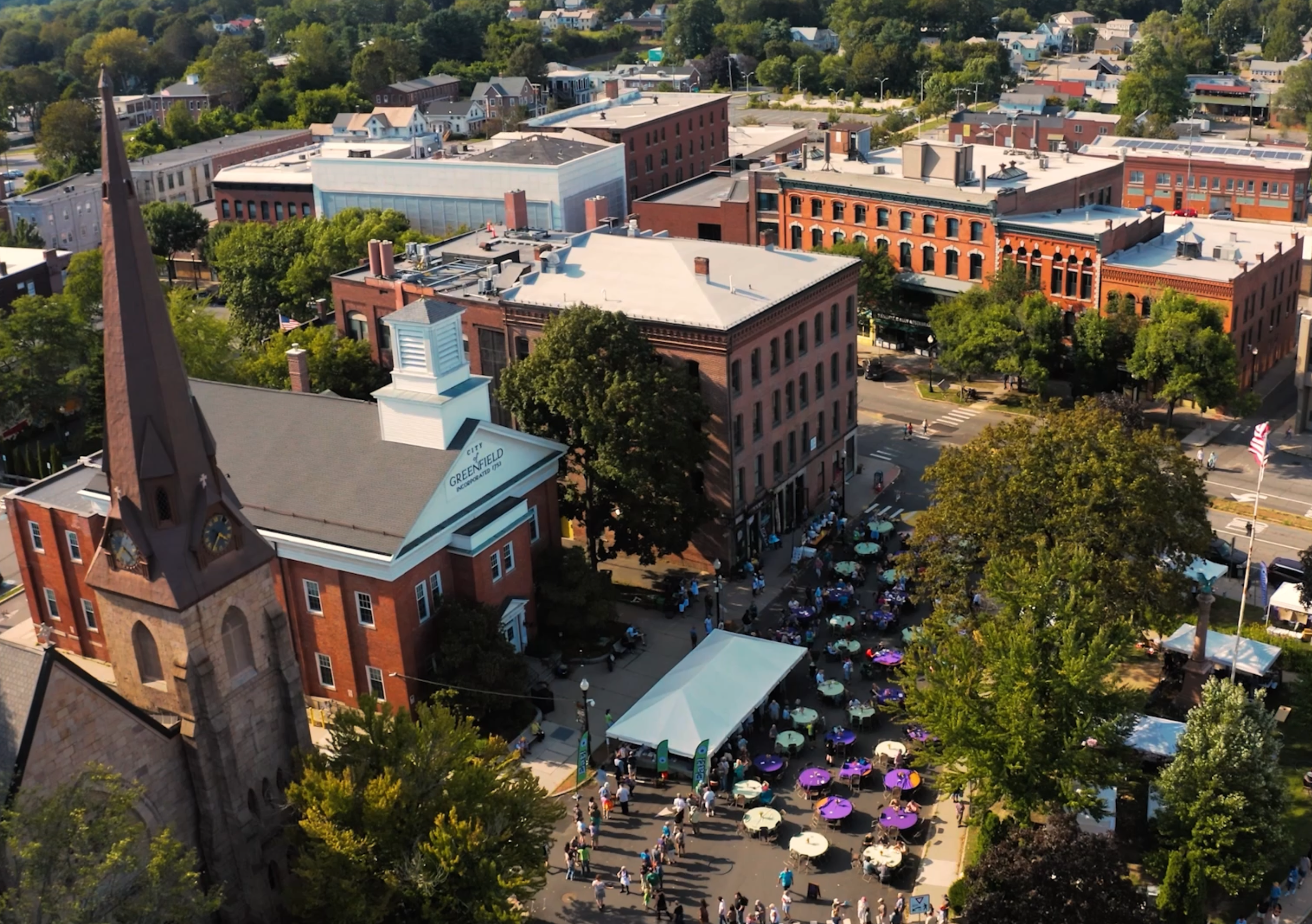 Aerial view of a town square filled with people walking, sitting under umbrellas, and gathering for an event on a sunny day, surrounded by historic brick buildings and green trees.