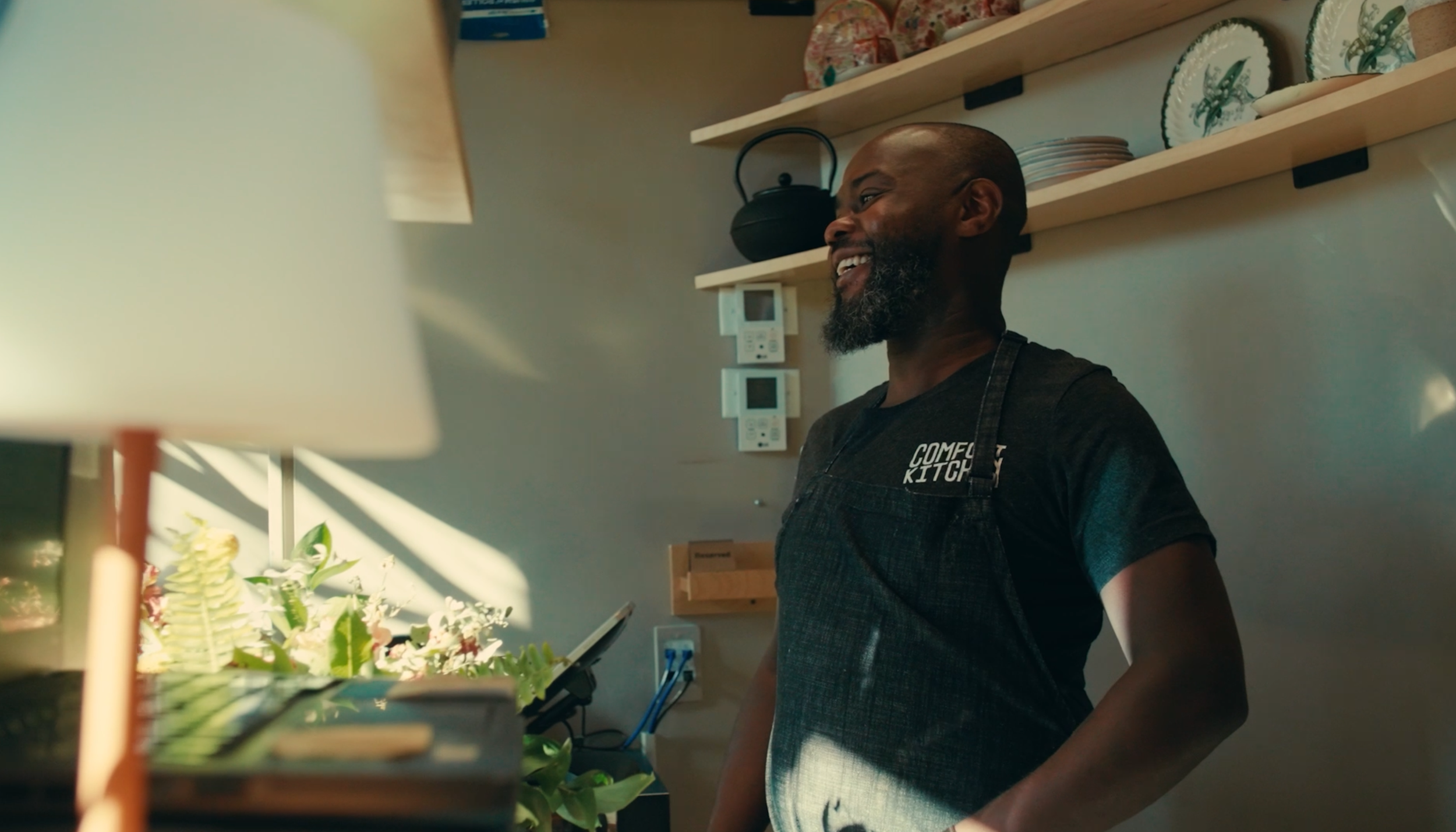 A smiling man with a beard wearing a black apron that says 'Comfort Kitchen' stands in a kitchen, with shelves holding plates and bowls behind him and a flower arrangement in the foreground.