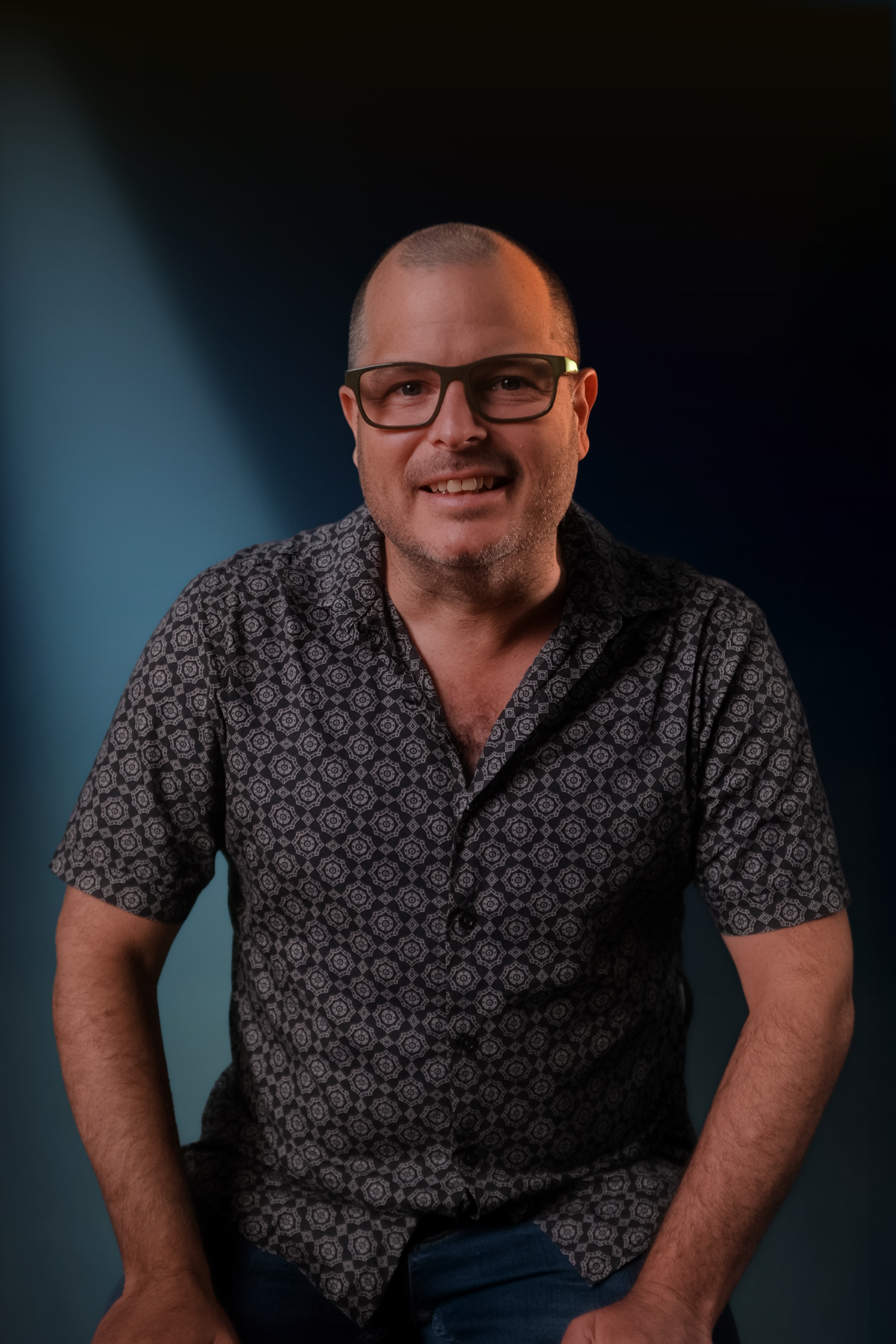 A man with glasses, short hair, and a patterned short-sleeved shirt smiling at the camera, seated against a dark gradient background.