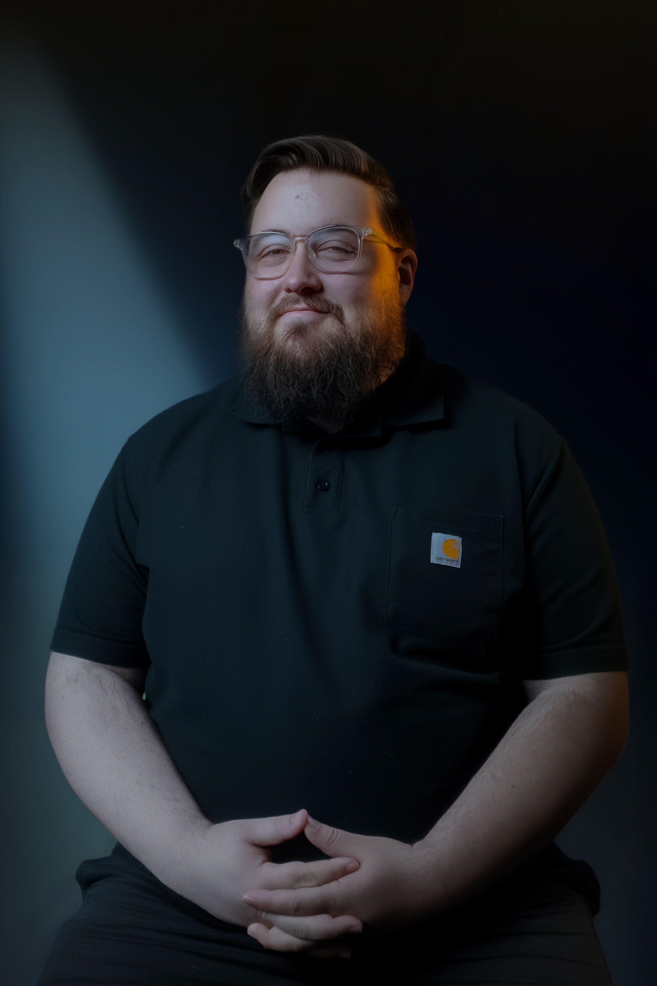 Portrait of a man with glasses and a beard, wearing a black Carhartt shirt, sitting with his hands clasped in his lap, against a dark background with a slight gradient.