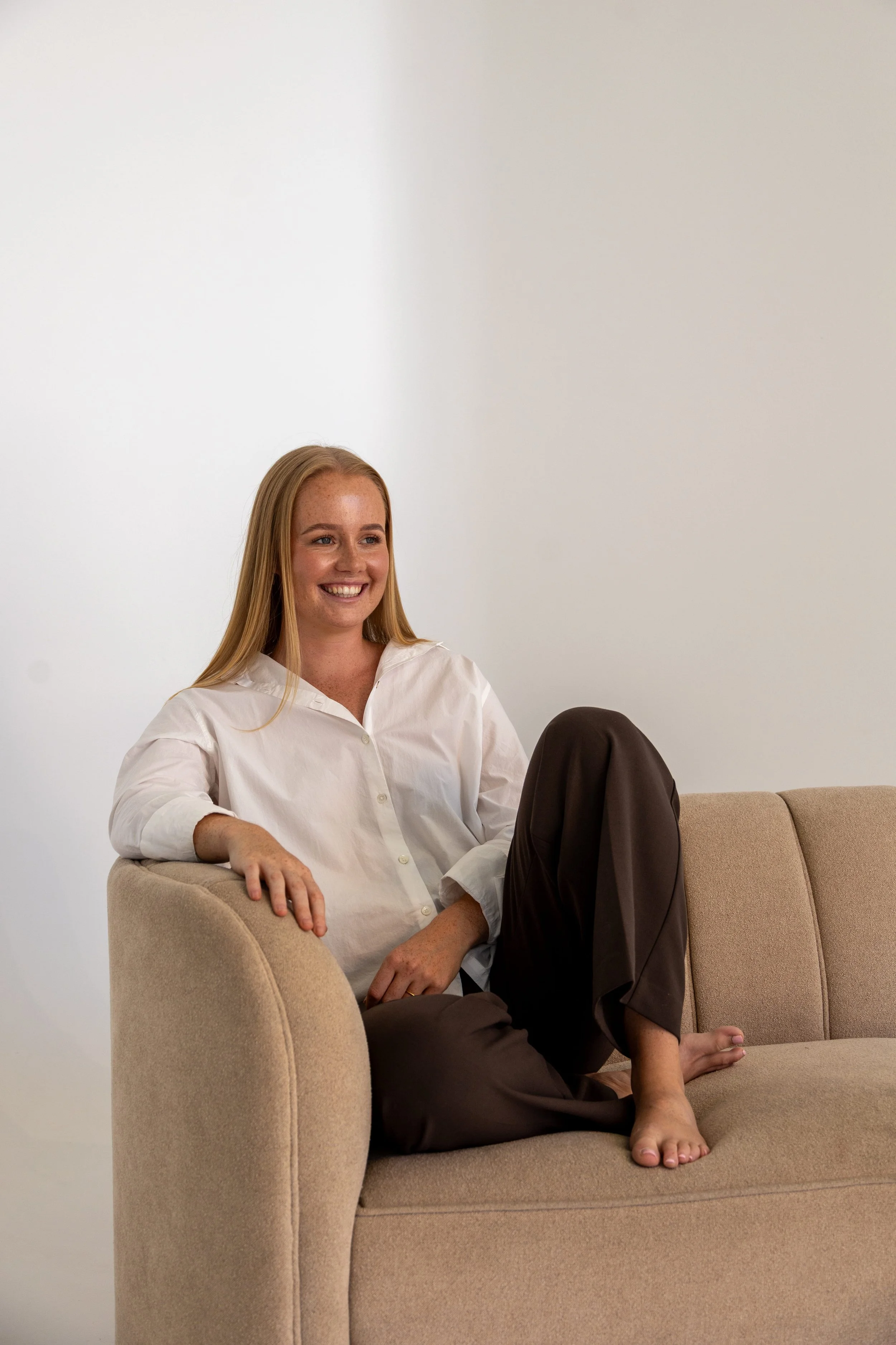 A young woman with long red hair and freckles sitting on a beige couch, smiling and looking to her right, with a plain white wall in the background.