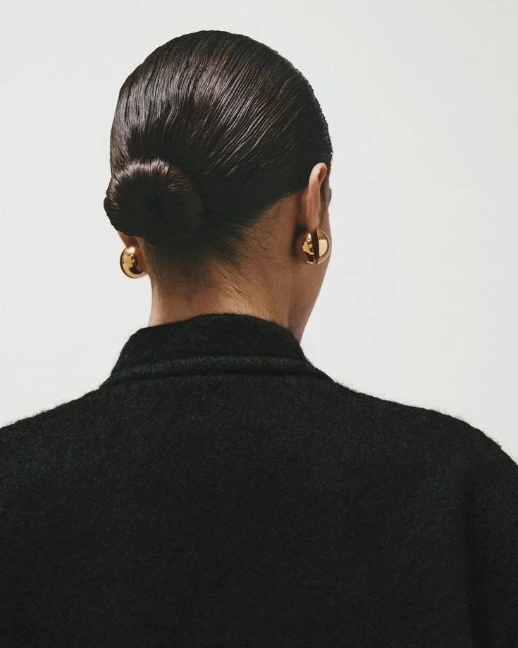 Back view of a woman with dark, slicked-back hair in a low bun, wearing gold hoop earrings and a black turtleneck against a plain white background.