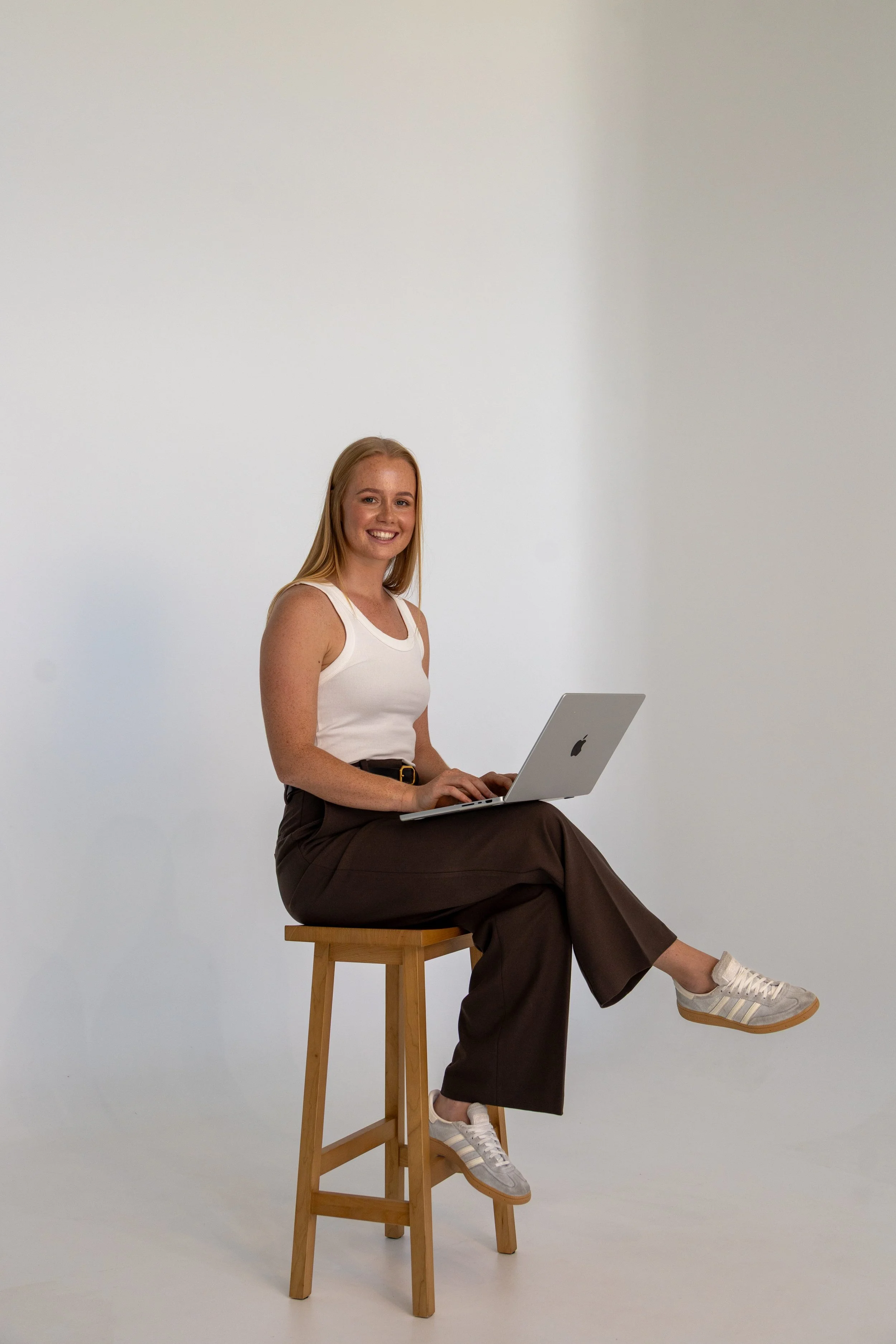 A smiling young woman with blonde hair, wearing a white tank top, brown wide-leg pants, and white sneakers, sitting on a tall wooden stool with one leg extended and resting on a lower rung, holding a silver MacBook laptop. The background is plain white.