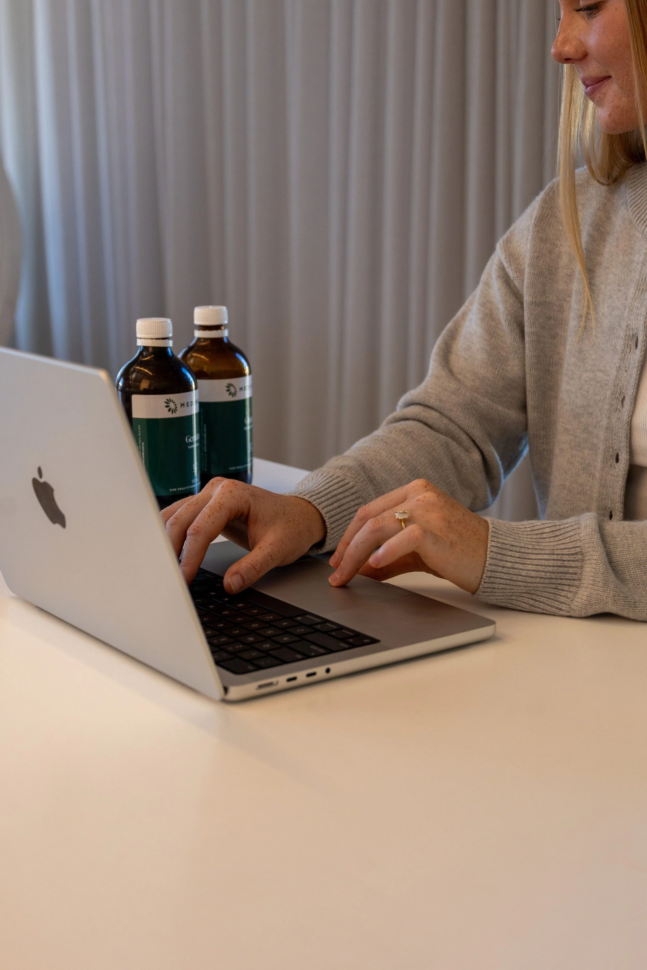 A woman in a beige sweater typing on a silver MacBook laptop at a white table, with two medication bottles on the table.