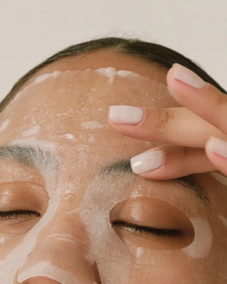 Close-up of a person with a facial sheet mask applied, eyes closed, person touching forehead with nails painted white.
