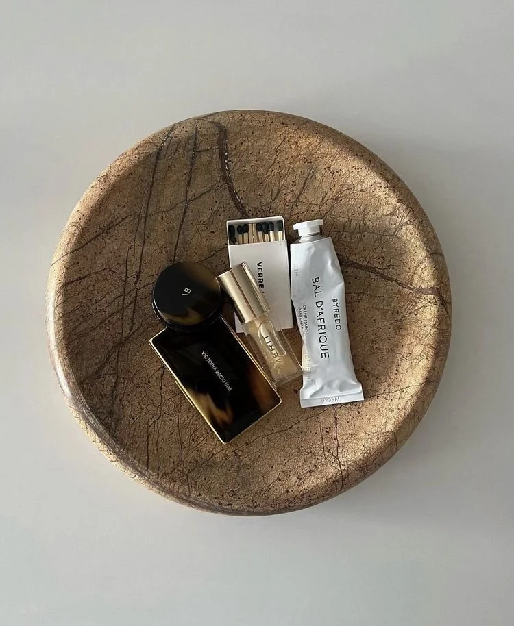 A round, textured, beige-colored dish with four perfume bottles and a small pack of disposable razors inside.