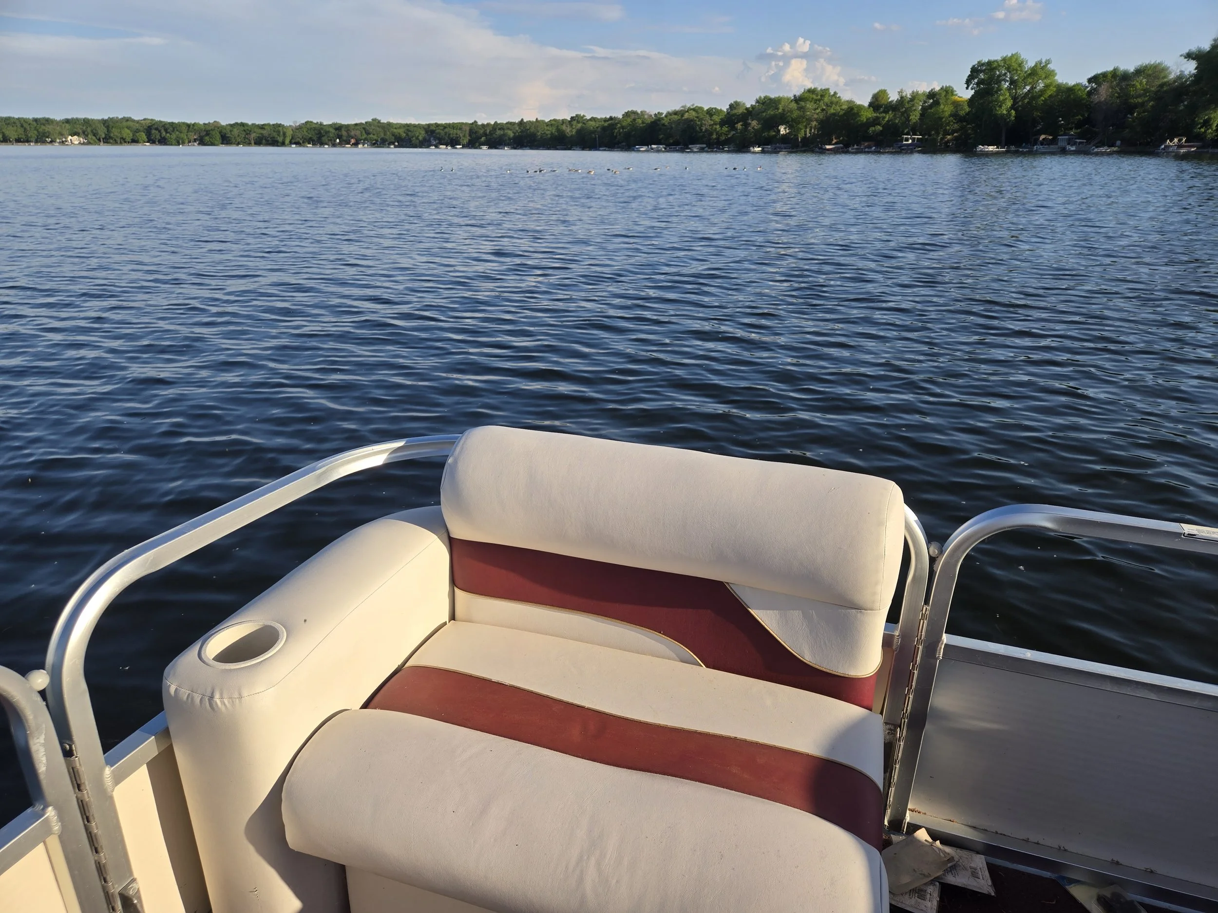 The photo shows the front of a boat with white and maroon seating. The boat is on a calm lake with a shoreline lined with trees and some houses. The sky is partly cloudy.