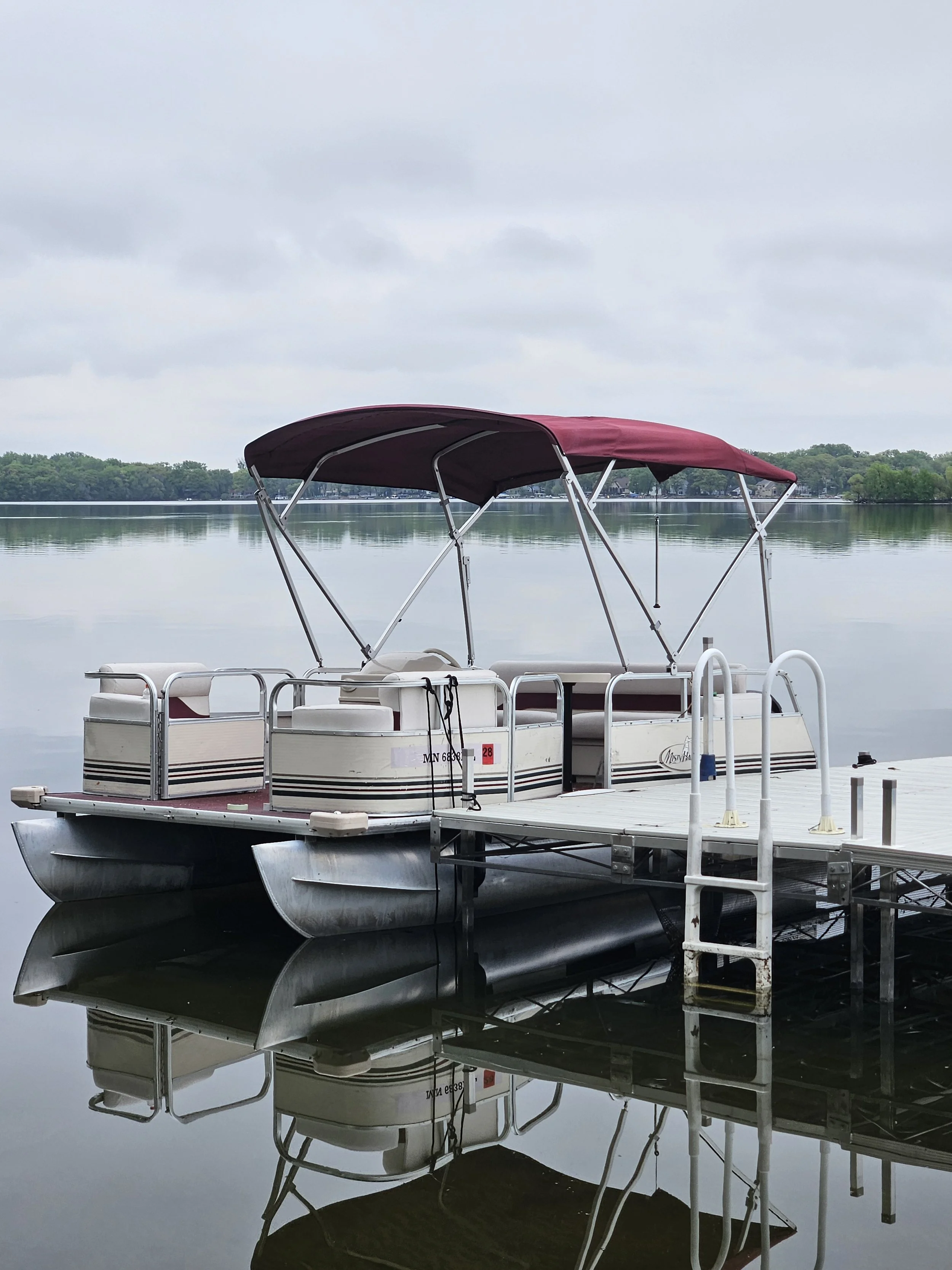 A pontoon boat with a maroon canopy docked at a pier on a calm lake, with a tree-lined shoreline in the background and reflections in the water.