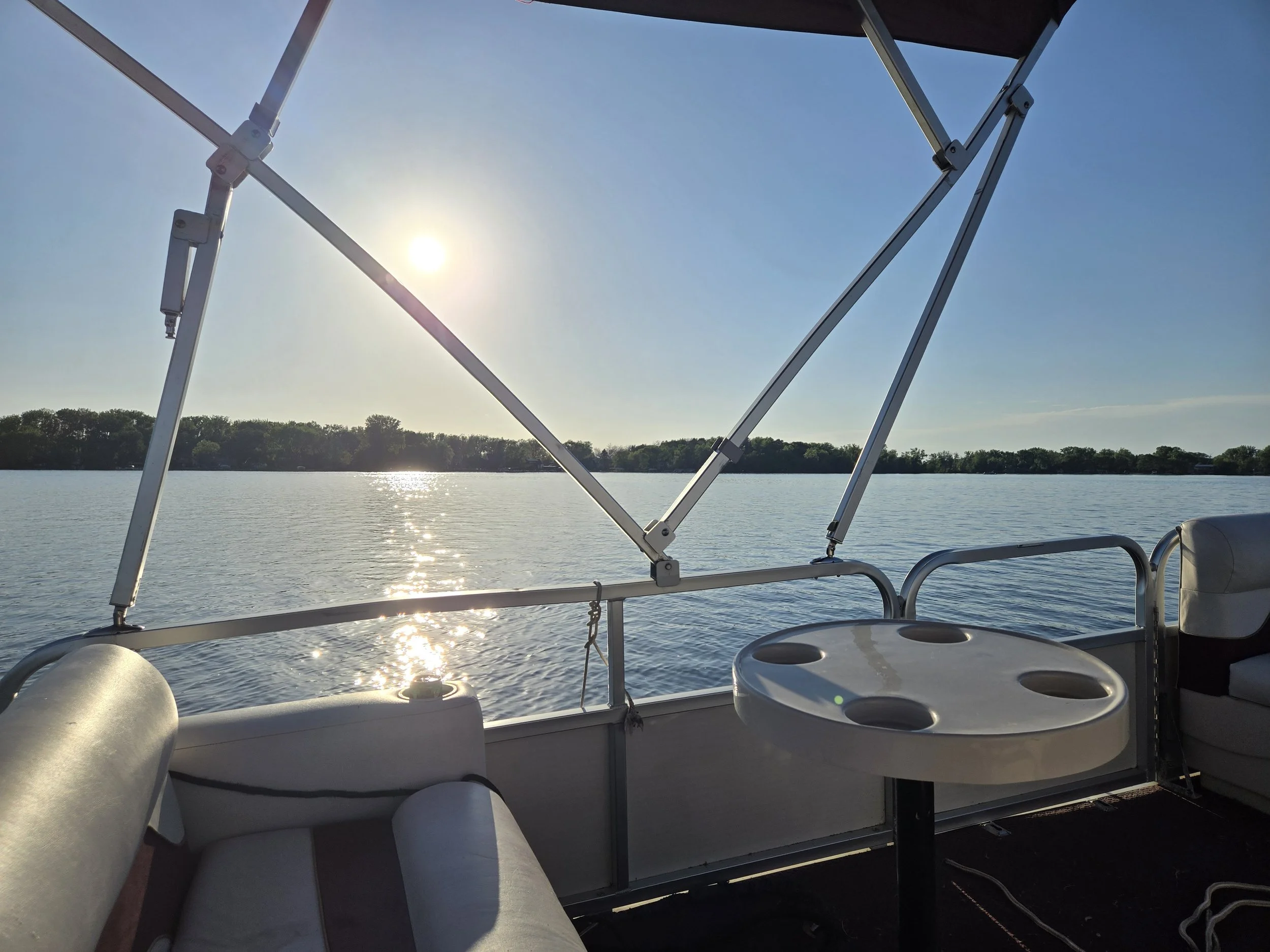 View from a boat's seating area showing white seats, a water body, and trees in the distance with the sun low in the sky.