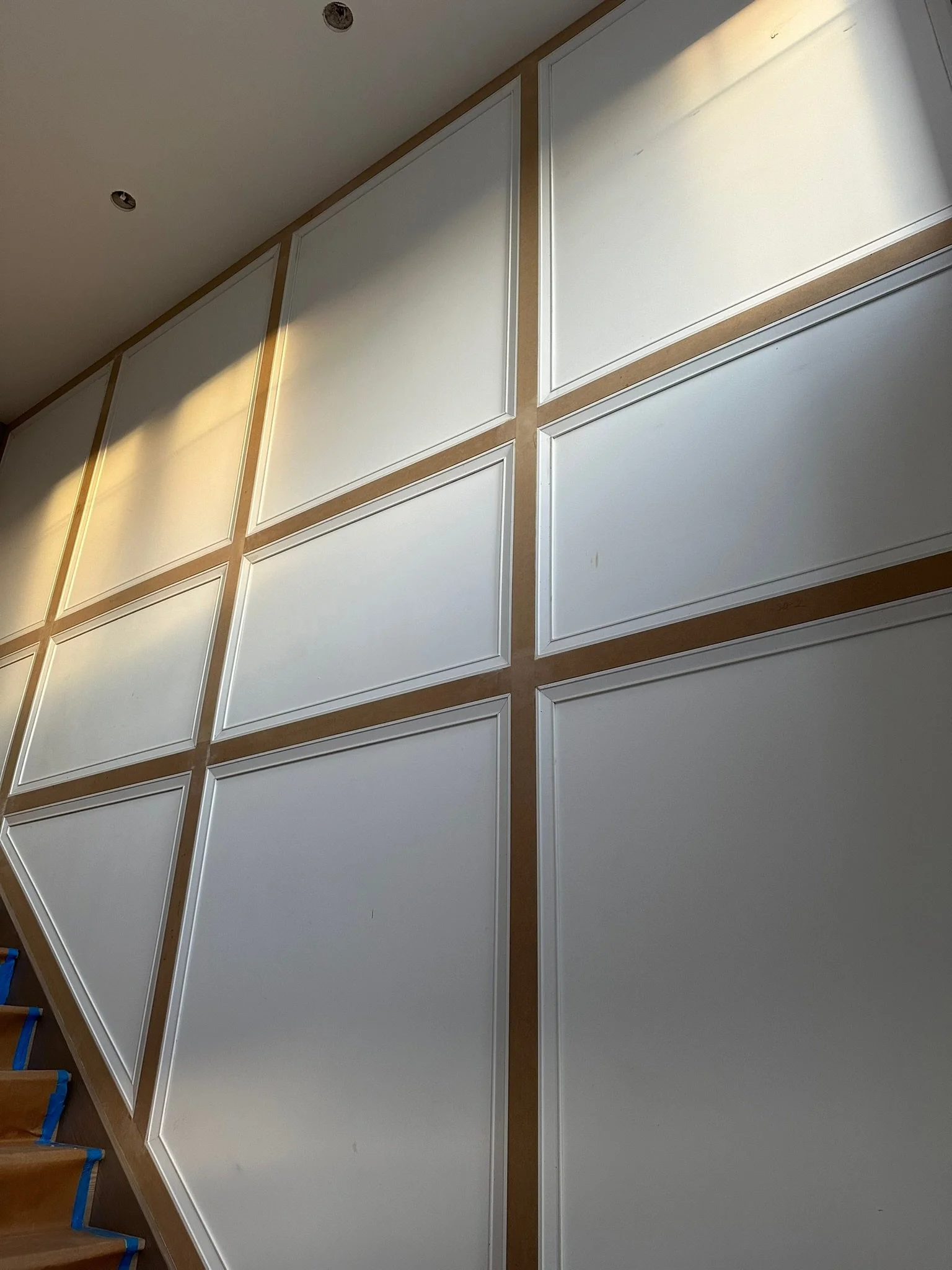 Interior room with white wall panels framed with wood, part of a staircase with blue painter's tape, and a ceiling with two recessed lights.