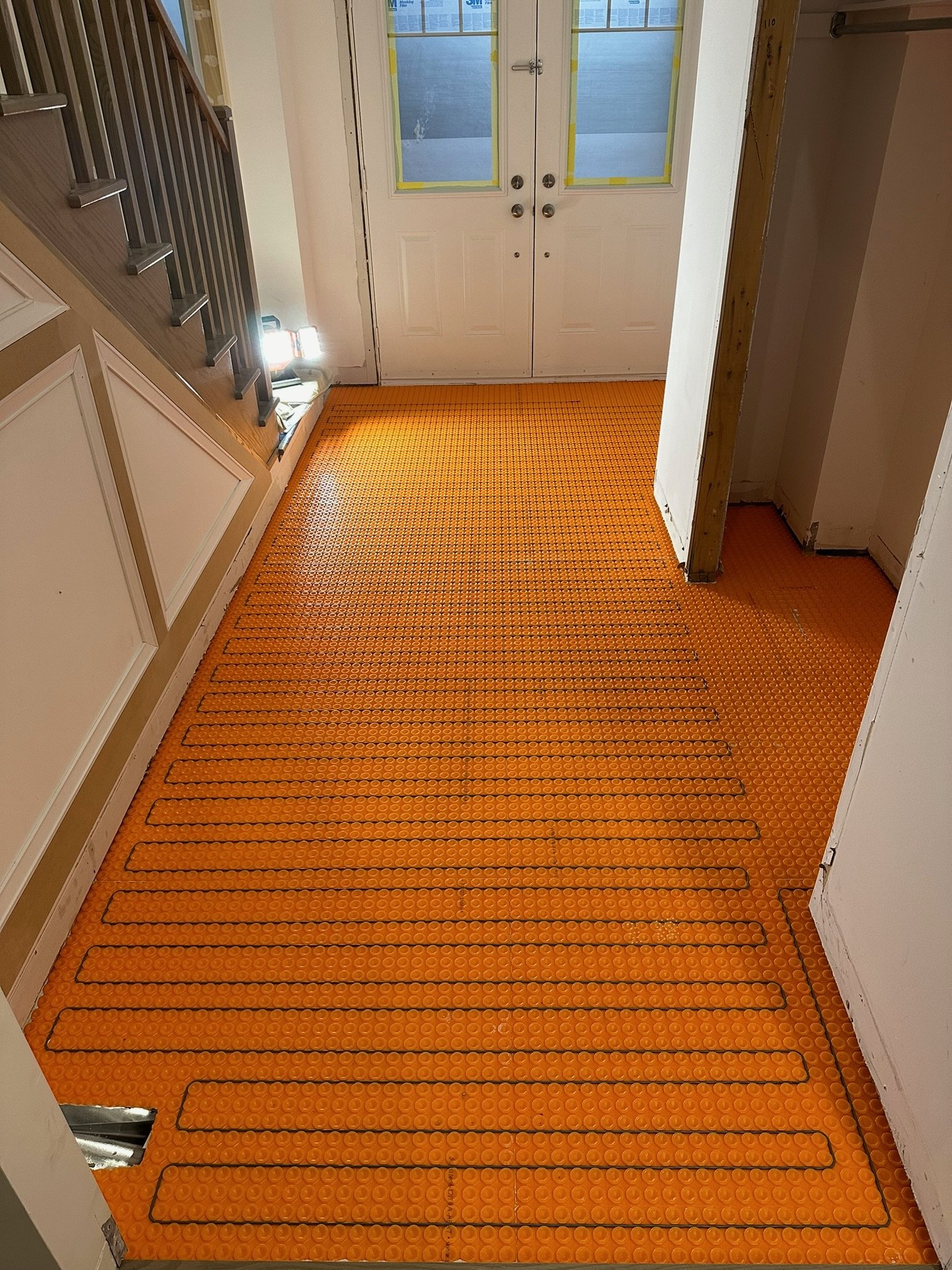 Interior view of an entryway with a bright orange rubber mat on the floor, partially beneath a staircase on the left and leading to a set of double doors with glass panels, which are taped for protection.