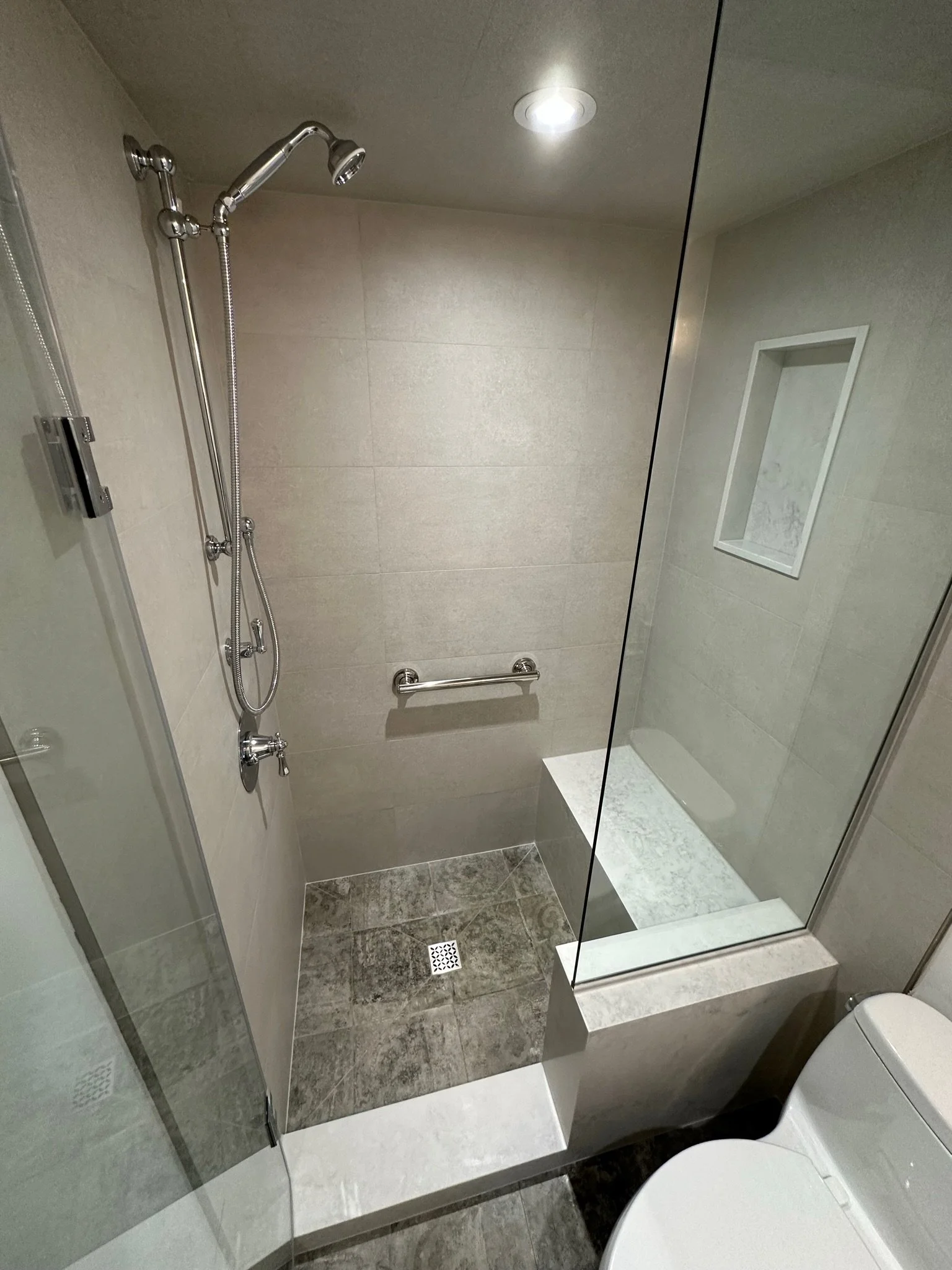 Modern bathroom shower area with a handheld showerhead, grab bar, recessed shelf, tiled walls, and a floor drain.