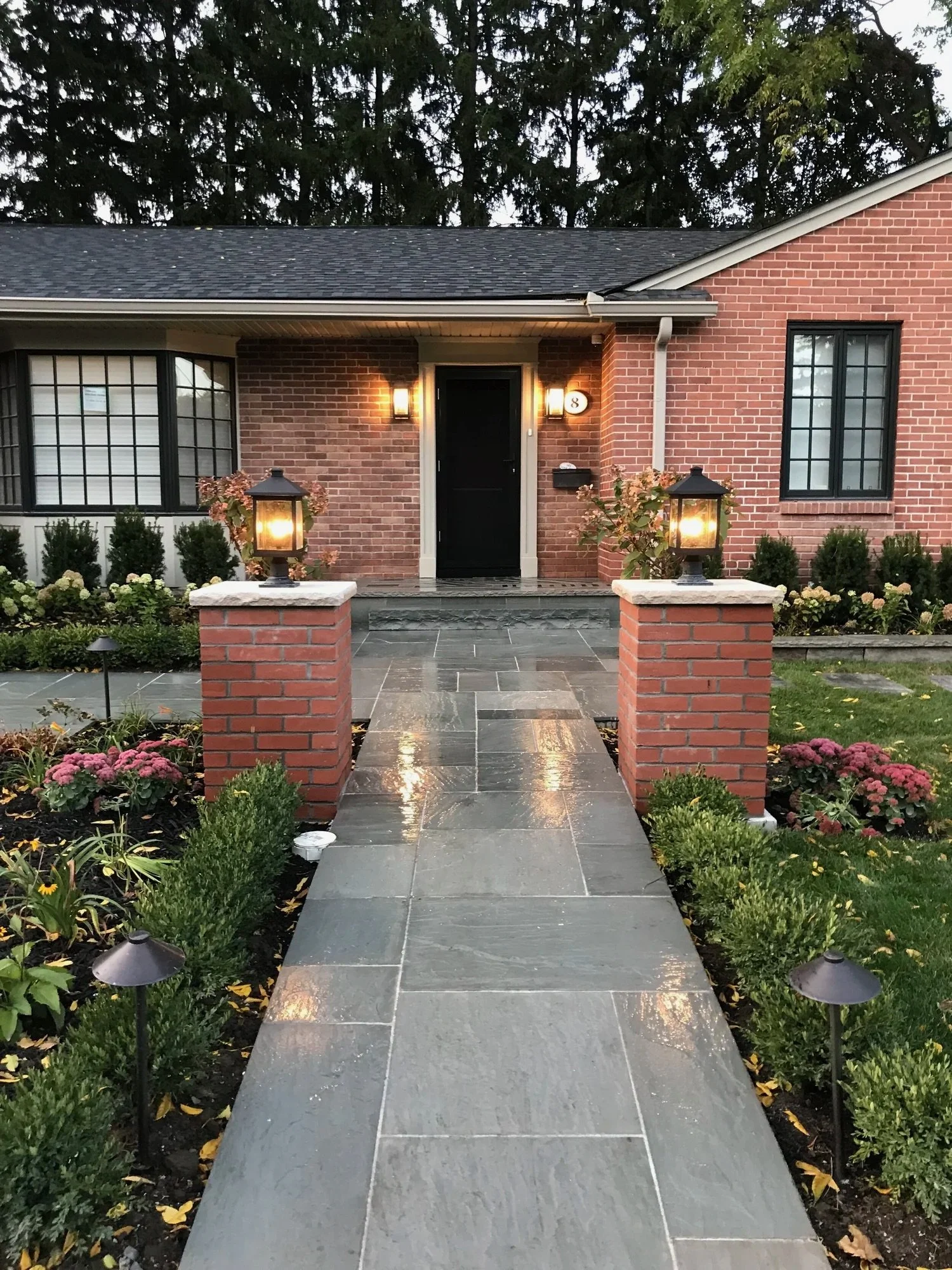 Landscaped yard in front of red brick home with two pillars with lanterns atop them flanking a beautifully laid stone walkway leading up to the porch. Custom lit walkway, beautiful brickwork and stone.