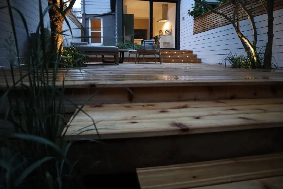 Outdoor wooden deck leading to a modern house interior with illuminated steps and outdoor furniture, surrounded by plants.
