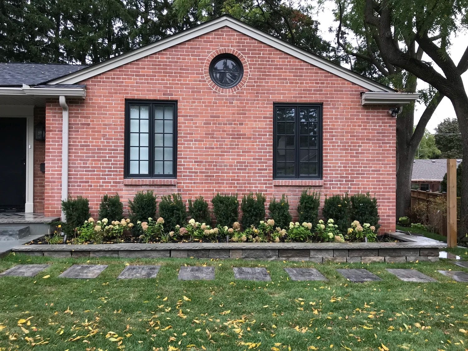 Red brick house with black-framed windows and a small circular window near the roof peak. Front yard with flower bed and green shrubbery, stone path, and a grassy lawn with fallen yellow leaves. Trees in background.
